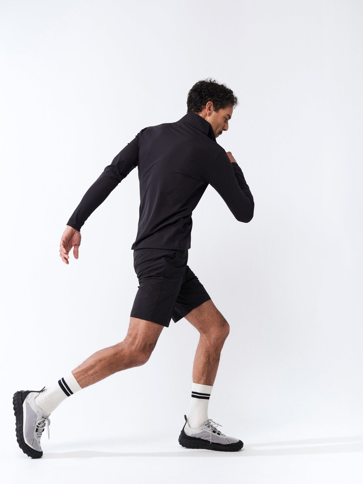 A person wearing a black jacket, black shorts, white socks, and sneakers strides forward energetically against a plain white background.