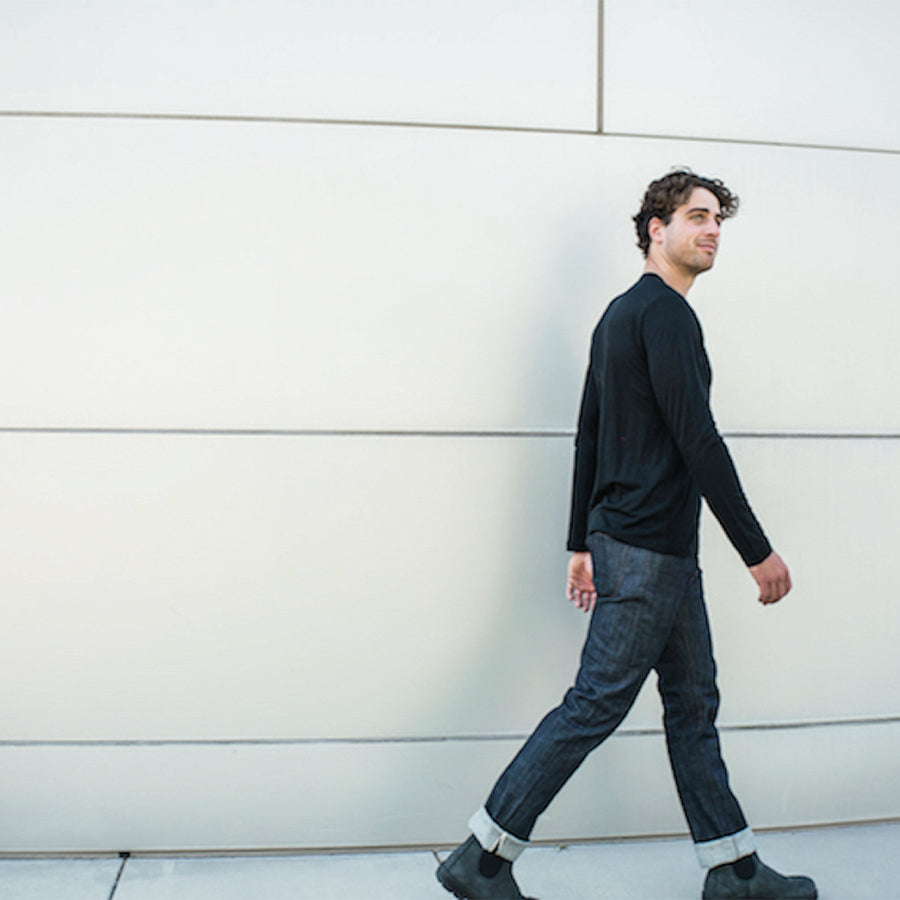 A man with curly dark hair, wearing a black long-sleeve shirt, cuffed jeans, and black boots, walks along a white, paneled wall. He smiles slightly while gazing to his left.
