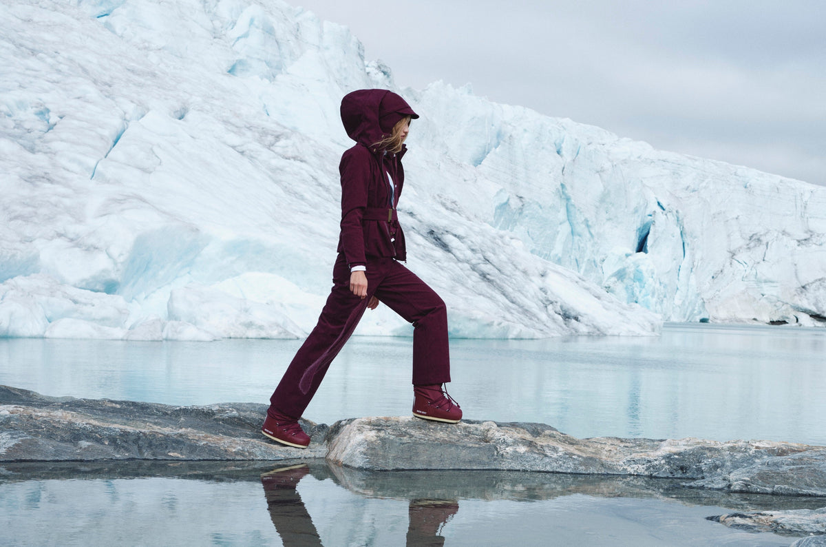 A woman in a burgundy hooded jumpsuit and matching winter boots steps onto a rock in front of a glacier and glacial lake.