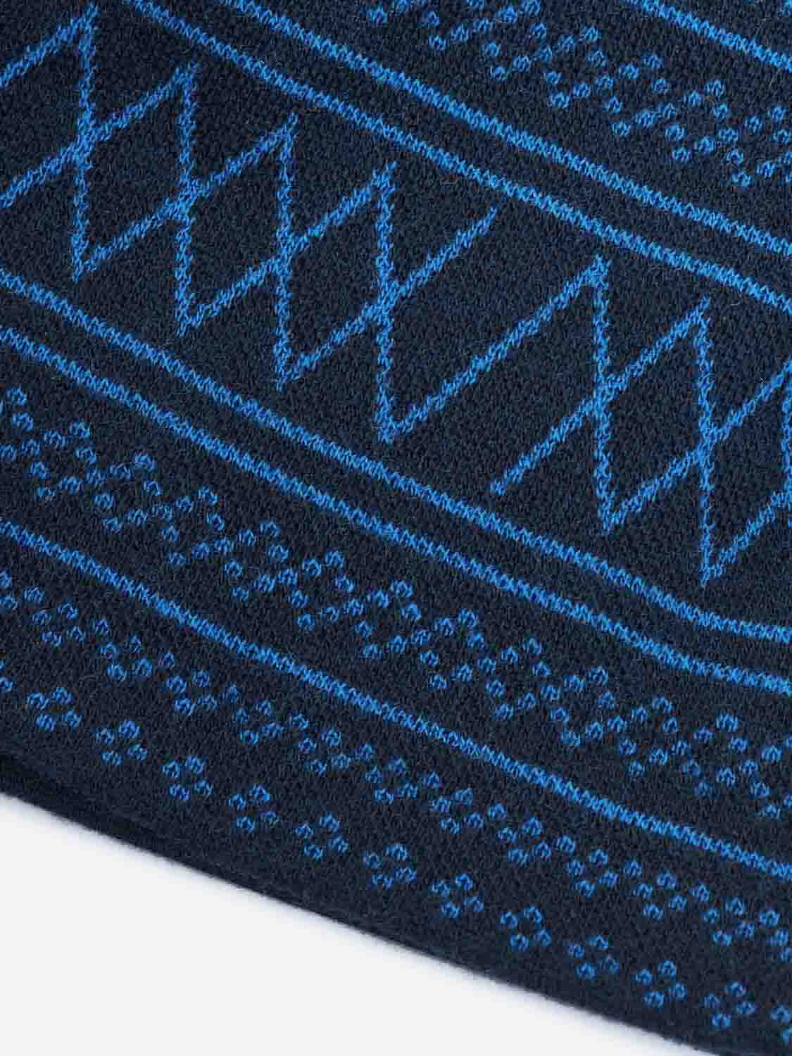 Close-up of a knitted jacquard fabric showcasing an intricate pattern of blue geometric zigzags and small dots against a dark backdrop, reflecting the traditional design of the We Norwegians Setesdal Hat Navy Blue.