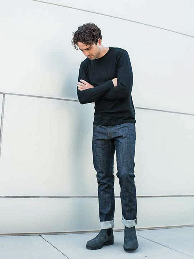 A person with curly hair, dressed in a slim fit We Norwegians Base One Longsleeve Men Black shirt, dark rolled-up jeans, and black boots, stands with arms crossed and head slightly bowed against a minimalist, light-colored wall backdrop.