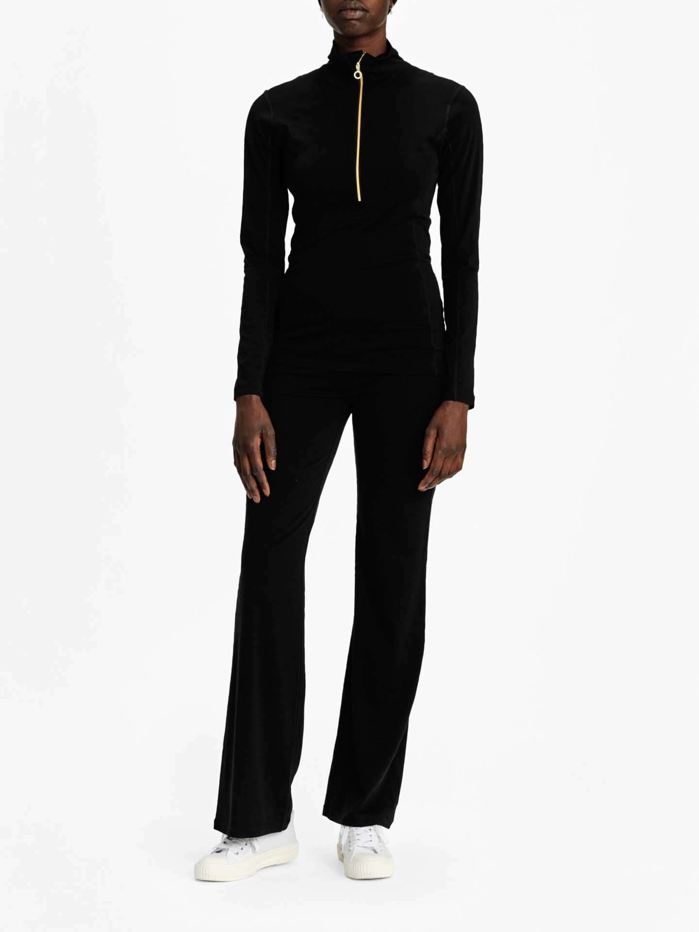 A person stands against a plain white background, wearing the Sno Merino Zip Up Women by We Norwegians, a fitted black long-sleeve top featuring a gold zipper running from the collar down to the chest, paired with matching black flared pants. They are also wearing white sneakers. The person's hands rest gently at their sides.