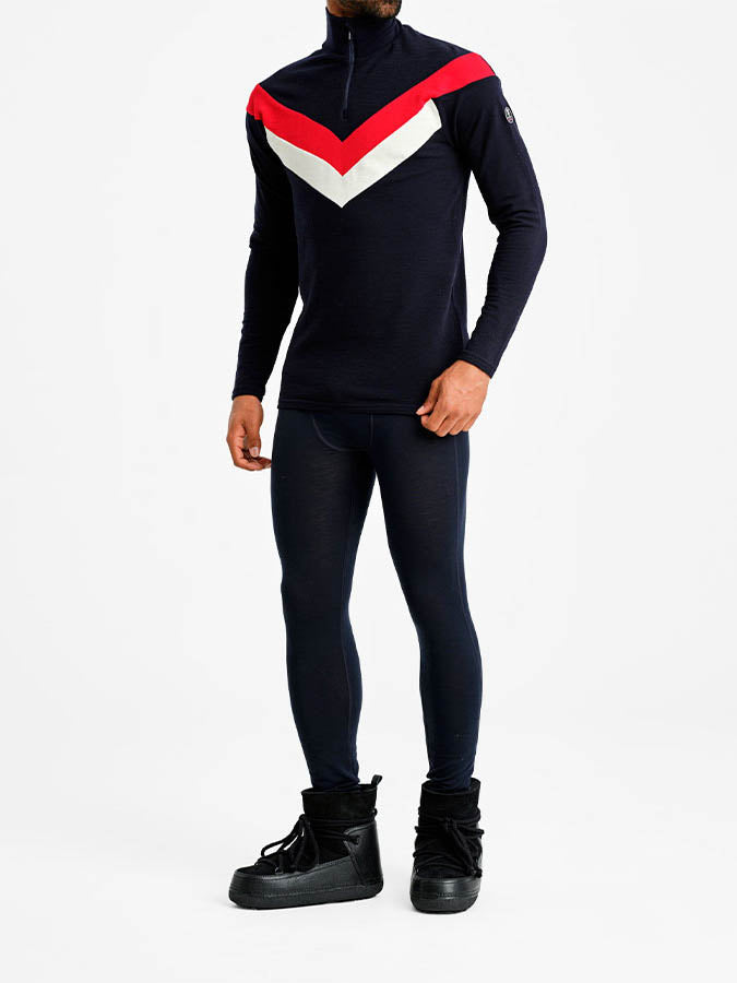 A person is dressed in We Norwegians' Voss Colblock 1/4 Zip Men Flag, a fitted navy blue outfit with color block detailing. The high-neck top features red and white chevron stripes across the chest. They are also wearing matching navy blue leggings and black snow boots, set against a plain white background.