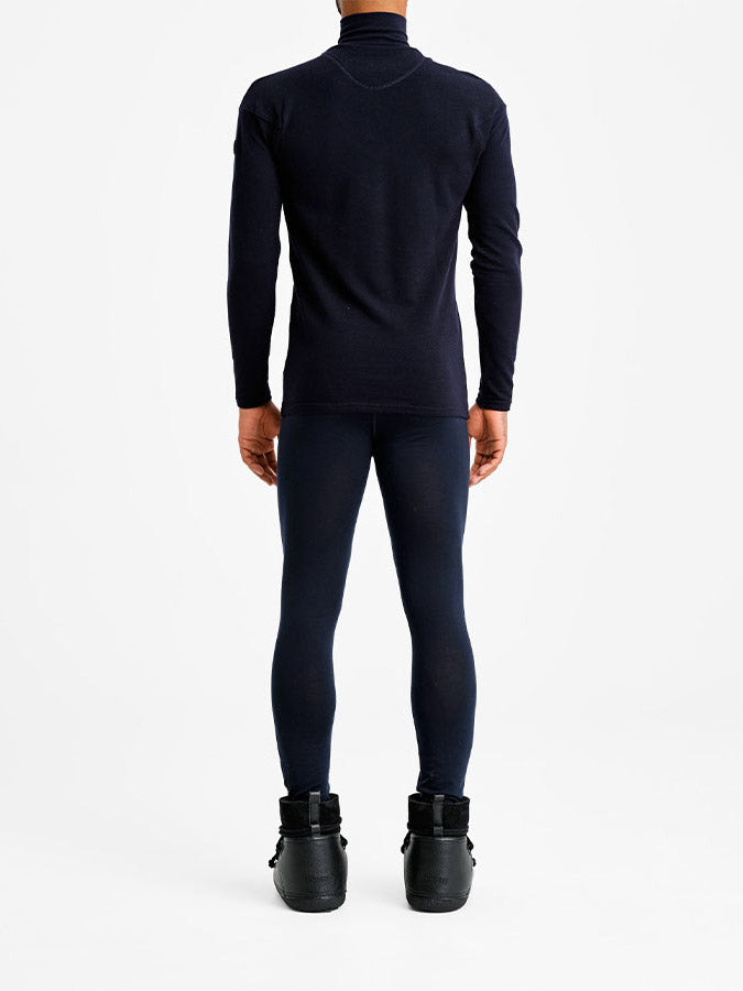A person stands facing away from the camera, dressed in a dark blue Voss Colblock 1/4 Zip Men Flag pullover and matching leggings by We Norwegians. They are wearing black shoes with a chunky sole. The background is plain white.
