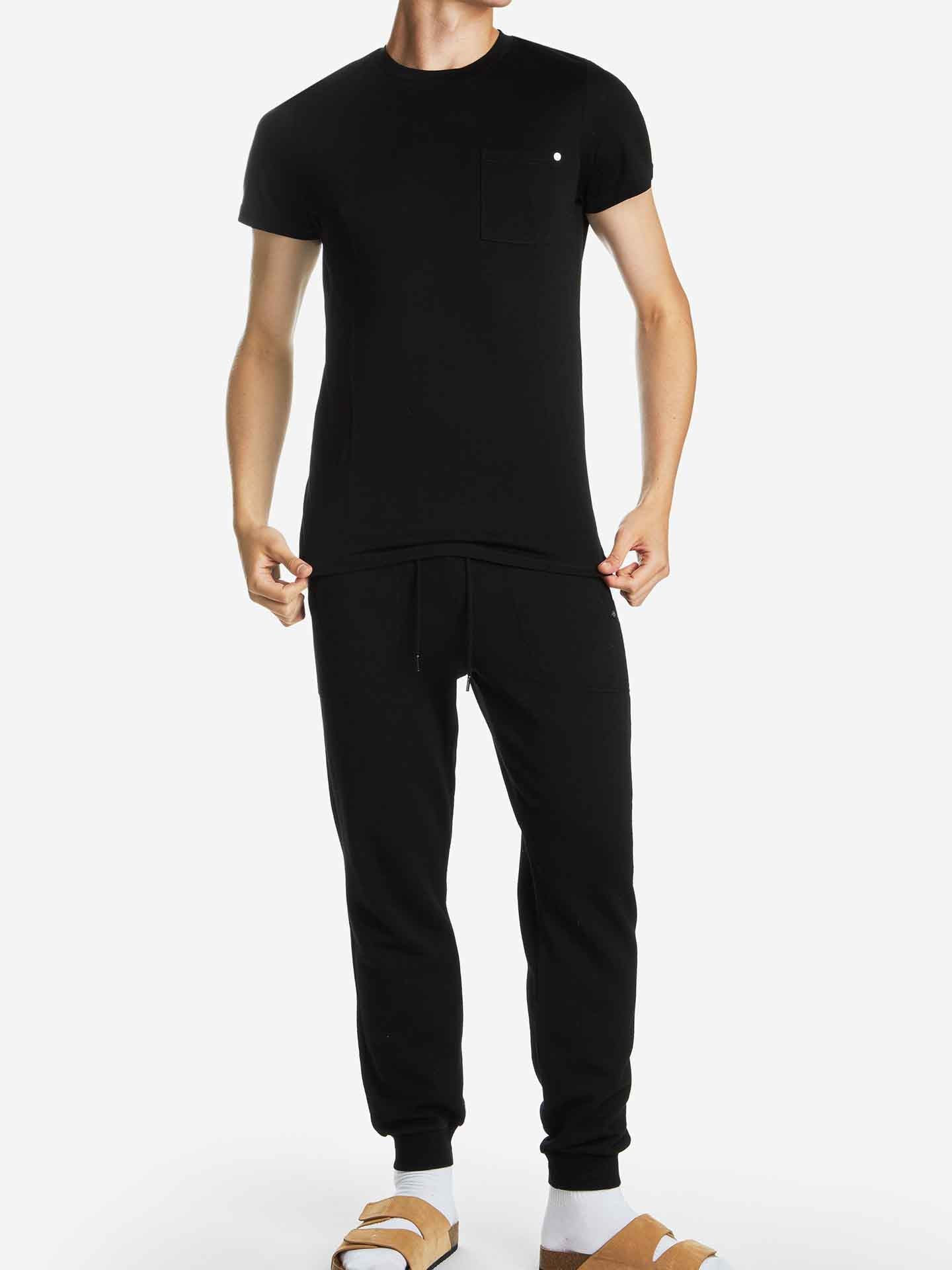 A person wearing the black Base One Pocket Tee Men by We Norwegians, featuring a classic crew neckline and a small chest patch pocket, stands against a plain white background. Paired with black jogger pants and beige slide sandals with white straps, their face is not visible.