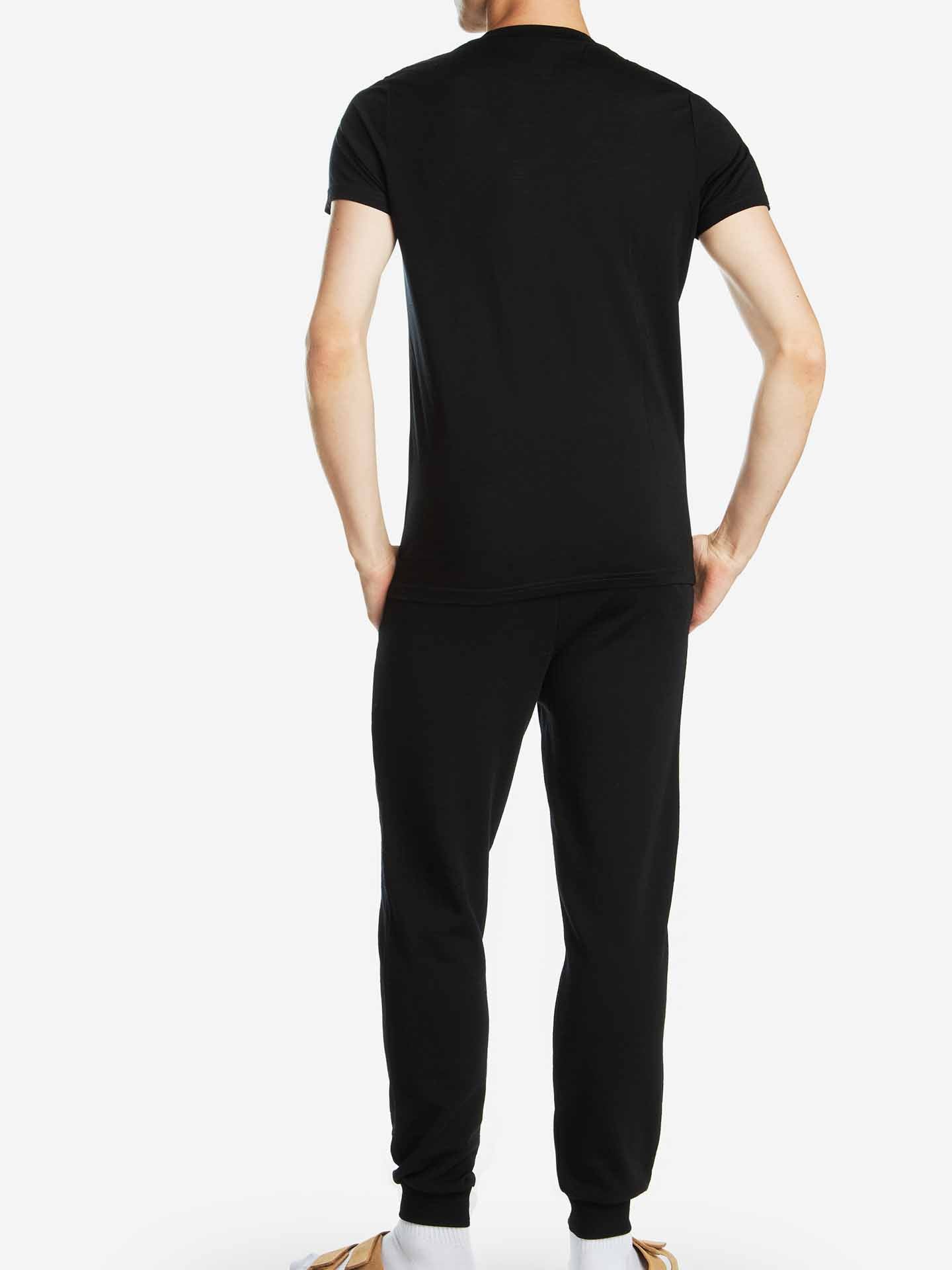 A person is standing with their back turned to the camera, wearing a We Norwegians Base One Pocket Tee Men in black, featuring a classic crew neckline and matching black pants. They are also wearing white socks and flip-flops. The background is plain white.