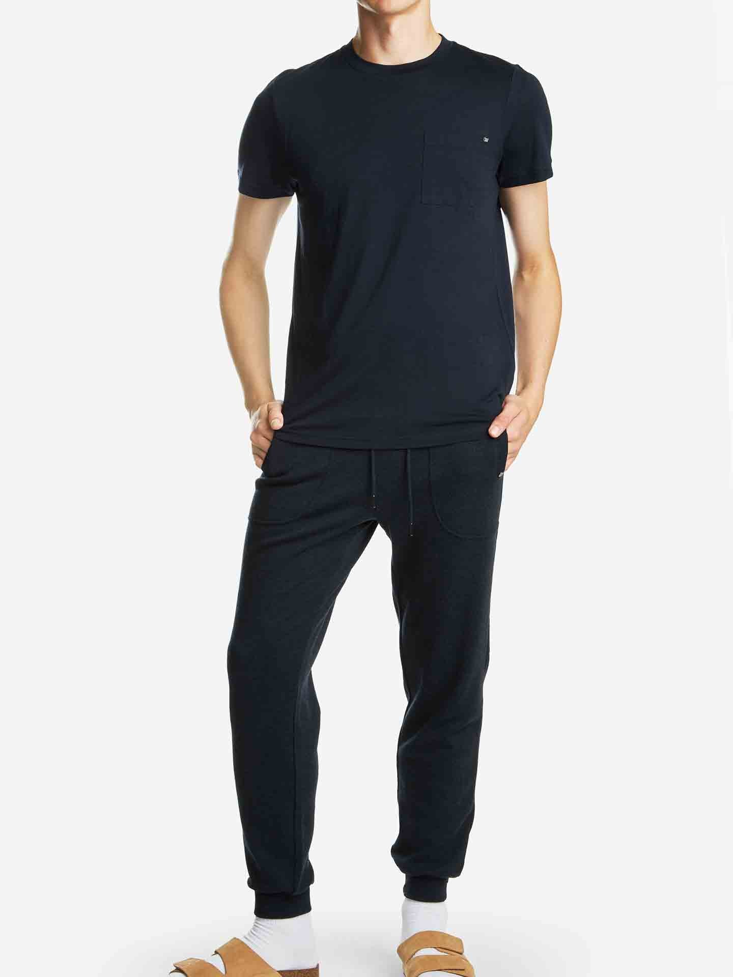 A person stands against a plain background, wearing a fitted navy t-shirt, identified as the "Base One Pocket Tee Men Navy" by We Norwegians, paired with black jogger pants and beige sandals over white socks. Their hands are in their pockets. The outfit exudes a casual and relaxed style. The head is not visible in the image.