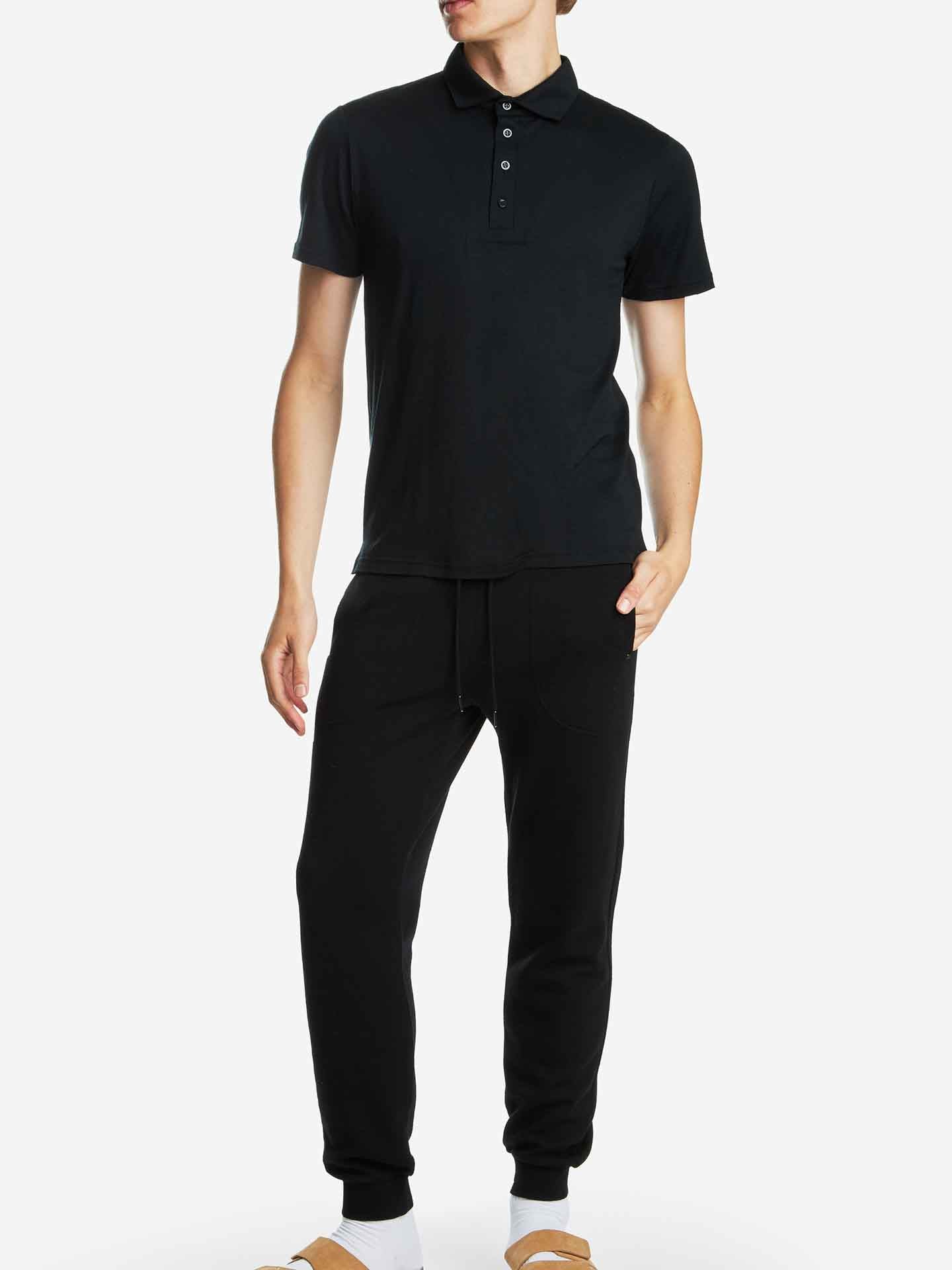 A person is standing against a plain background, wearing a We Norwegians Base One Polo Men Black and black jogger pants. The individual is also wearing white socks and light brown sandals. One hand is resting in their pants pocket while the other hangs by their side.
