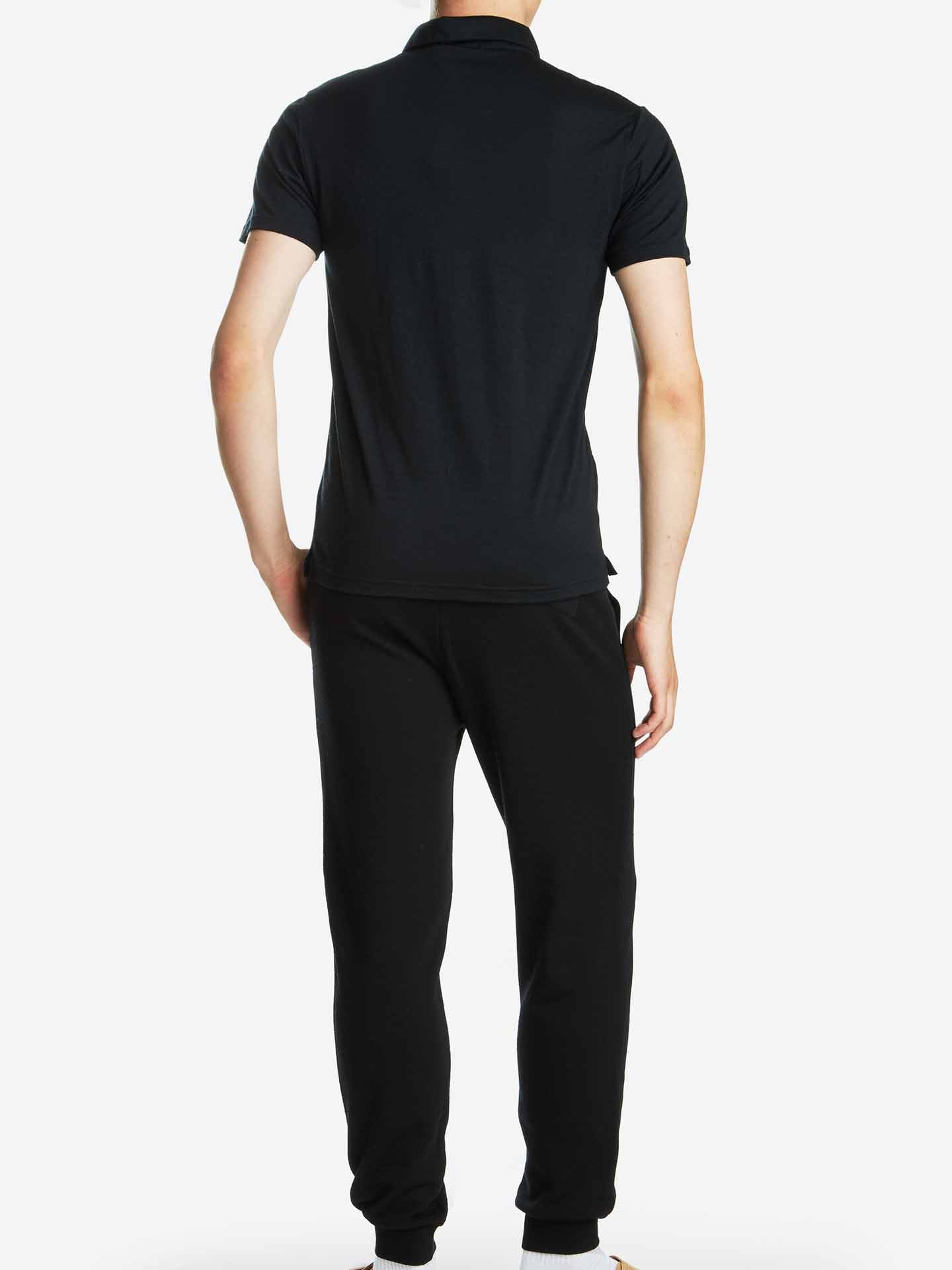 A person wearing the We Norwegians Base One Polo Men Black, made of super fine merino wool, and black pants stands facing away from the camera against a white background. The individual's arms are relaxed by their sides.