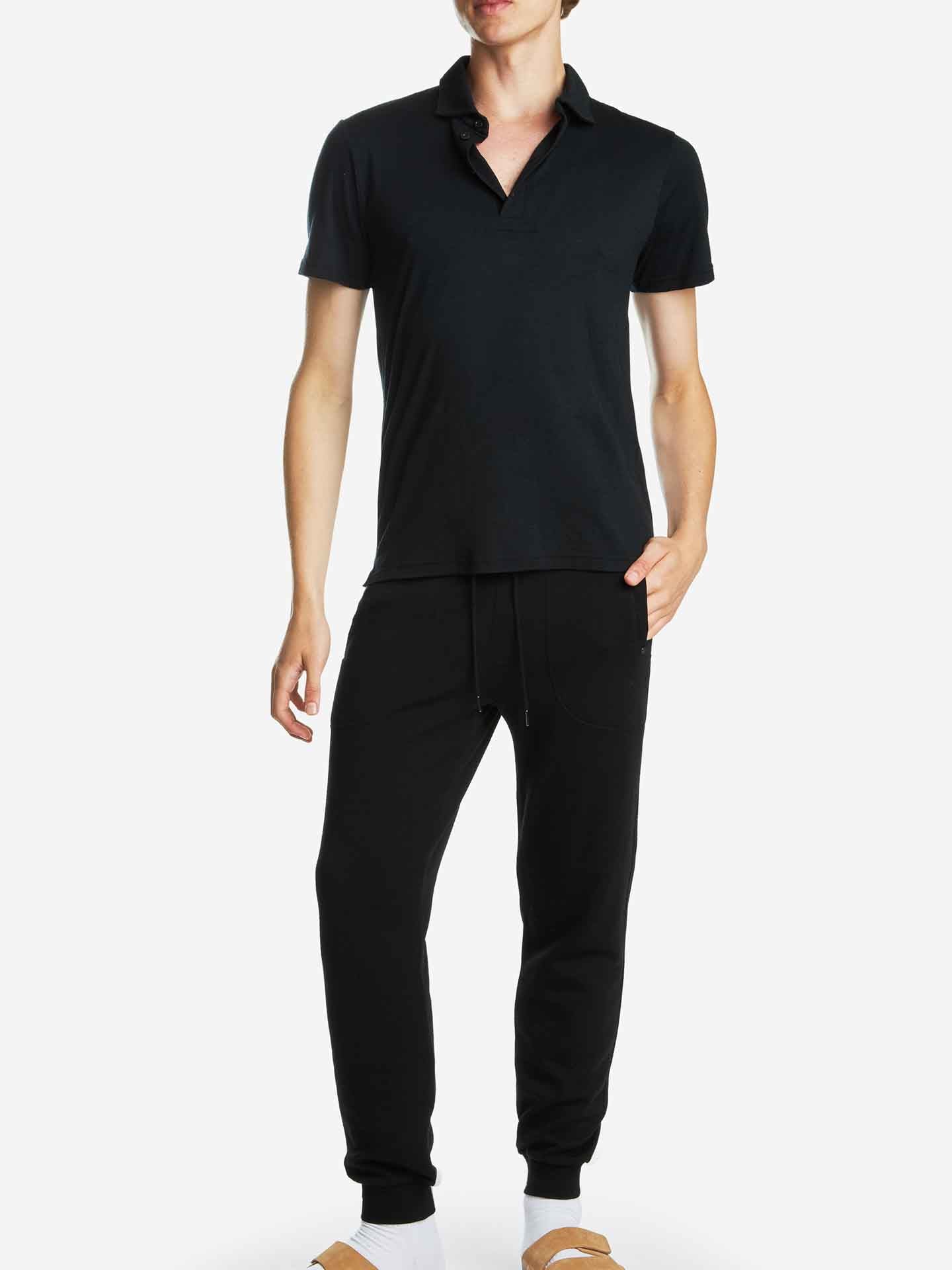 A person is wearing a short-sleeved Base One Polo Men Black shirt by We Norwegians, along with black jogger pants and white socks paired with brown slip-on footwear. They are standing with one hand in their pocket against a plain white background.