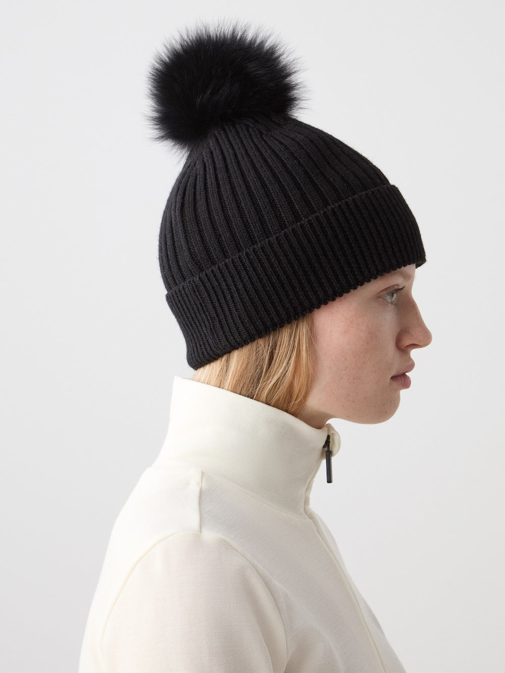 A woman in profile wears the We Norwegians Hemsedal Hat Women—a black ribbed knit luxury hat with a large black faux fur pom-pom—and a white high-collar jacket, capturing Scandinavian winter style against a plain, light background.