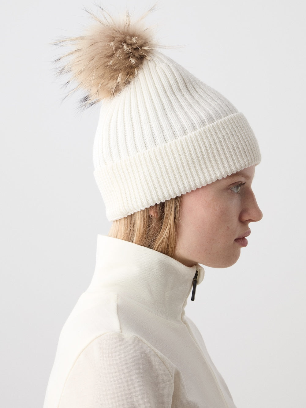A woman in a cream zip-up jacket wears the We Norwegians Hemsedal Hat—a luxury white ribbed knit beanie with a large, fluffy, light brown pom-pom—shown in profile against a plain light background.