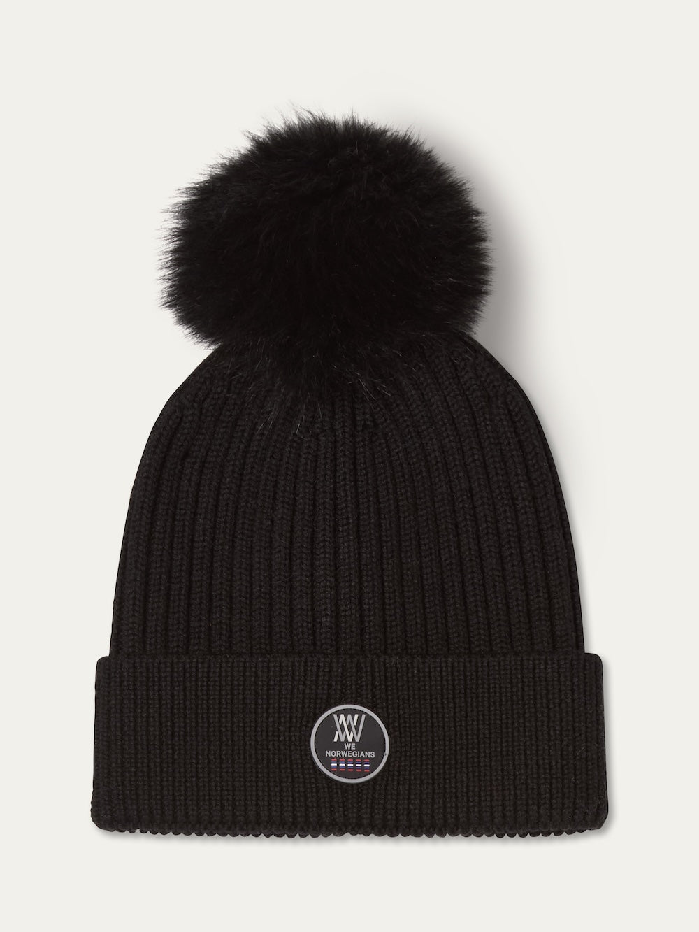 The We Norwegians Hemsedal Hat Women is a black ribbed knit beanie styled after Scandinavian winter accessories, featuring a large fluffy pom-pom and a folded brim with a circular patch logo on the front.