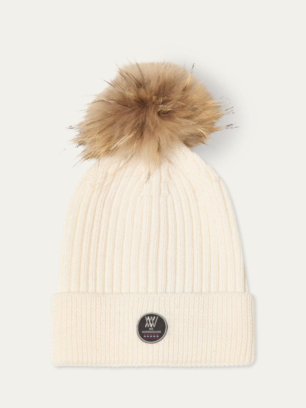 The Hemsedal Hat Women by We Norwegians is a cream ribbed knit beanie with a large brown faux fur pom-pom and a black circular logo patch on the front cuff, inspired by Scandinavian winter accessories.