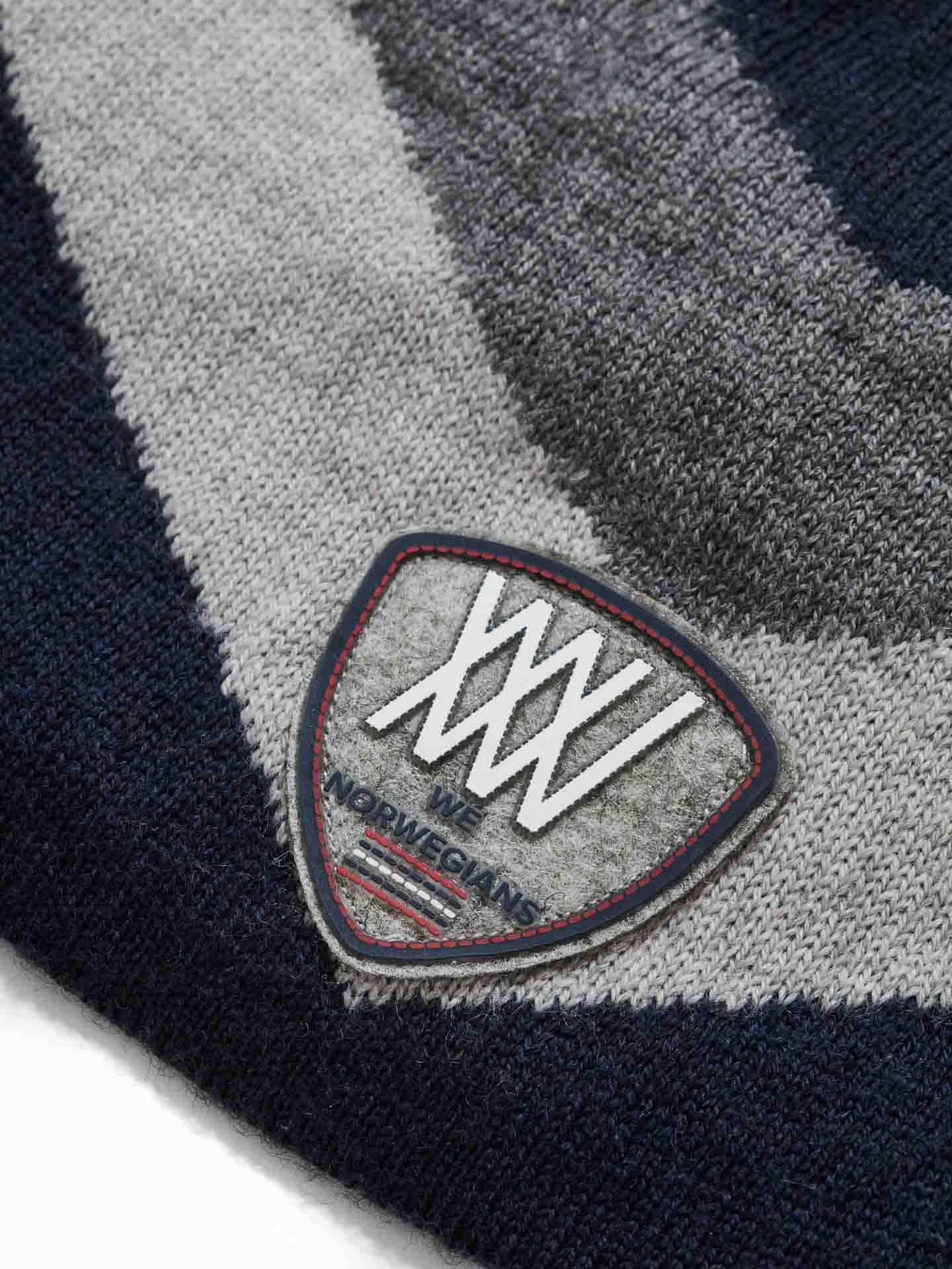 Close-up image of a patch sewn onto the Beito Hat. The shield-shaped patch prominently displays the Roman numerals "XXXVII," with "WE NORWEGIANS" text below. The Merino wool fabric, known for its breathability, features a pattern in shades of grey and dark blue.