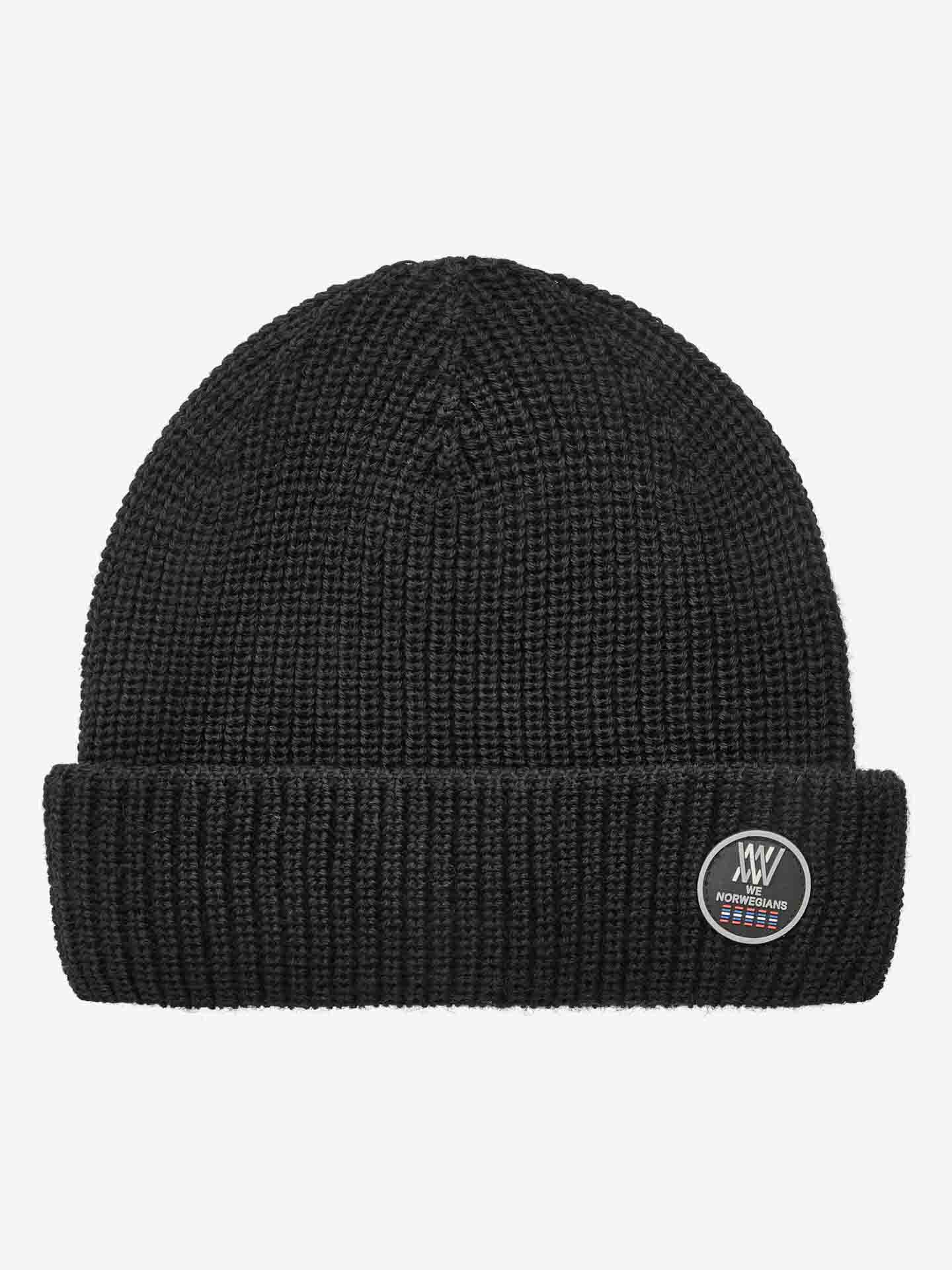 The Kvitfjell Hat by We Norwegians is a black ribbed knit beanie featuring a folded cuff adorned with a round patch displaying the text "XXV Norwegian" and a small flag symbol. Made from breathable Merino wool, this hat boasts a simple, classic design with a snug fit and natural antibacterial properties.