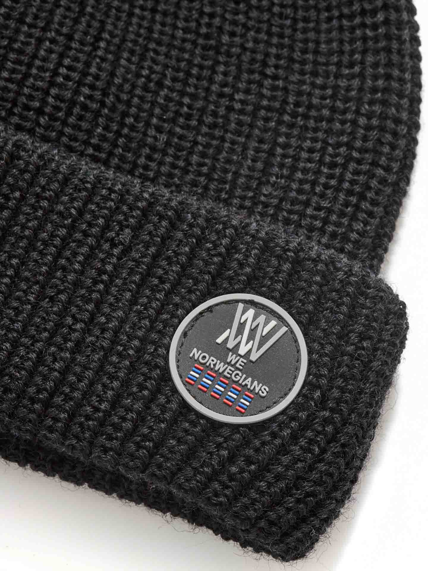 Close-up image of a black, knitted Kvitfjell Hat by We Norwegians featuring a circular patch. The patch includes a logo with a stylized "W" and "N," the words "We Norwegians," and a small flag design at the bottom. The beanie’s texture, known for its antibacterial and breathable properties, is clearly visible.