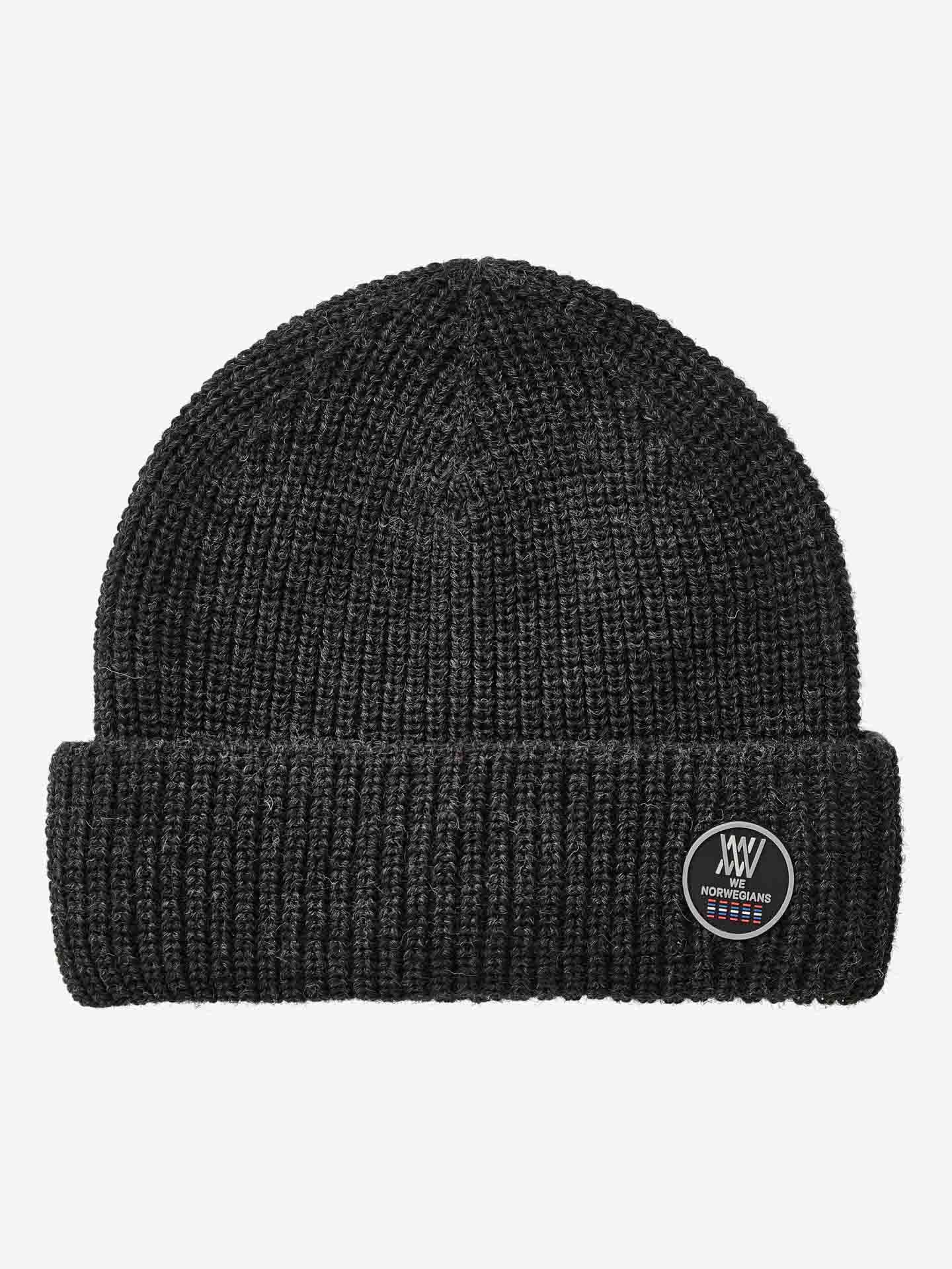 Close-up image of the Kvitfjell Hat, a dark gray, ribbed knit beanie from We Norwegians. Made from breathable Merino wool with a folded brim, it features a small circular patch with a logo that includes text and graphic design. The beanie is set against a plain white background.