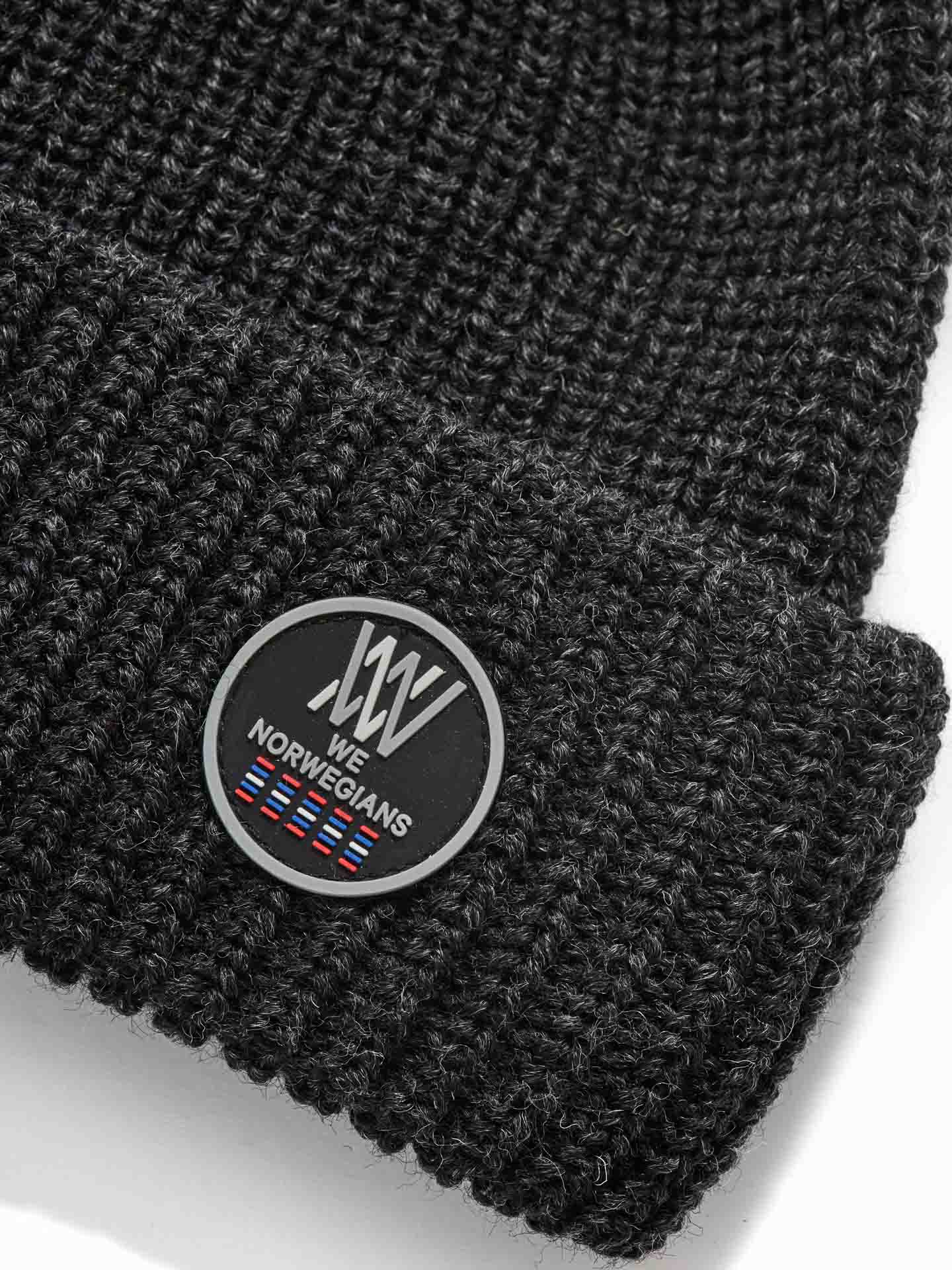 Close-up of the Kvitfjell Hat, a dark gray knitted beanie made from breathable Merino wool. It features a circular patch on the folded brim displaying the We Norwegians logo, text "We Norwegians," and a series of small multicolored flags at the bottom. The texture of the knit is clearly visible and naturally antibacterial.