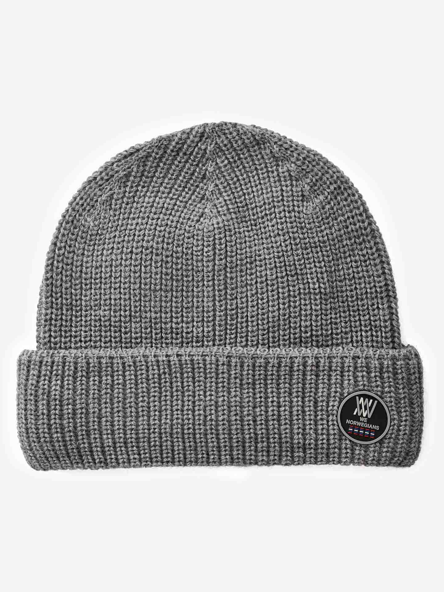A Kvitfjell Hat by We Norwegians, this gray knit beanie features a ribbed texture and is crafted from breathable Merino wool. It has a fold-over cuff adorned with an oval black patch displaying a white logo, with the words "HONEYBEARD & CO." written in white above the logo. The beanie is showcased against a plain white background.