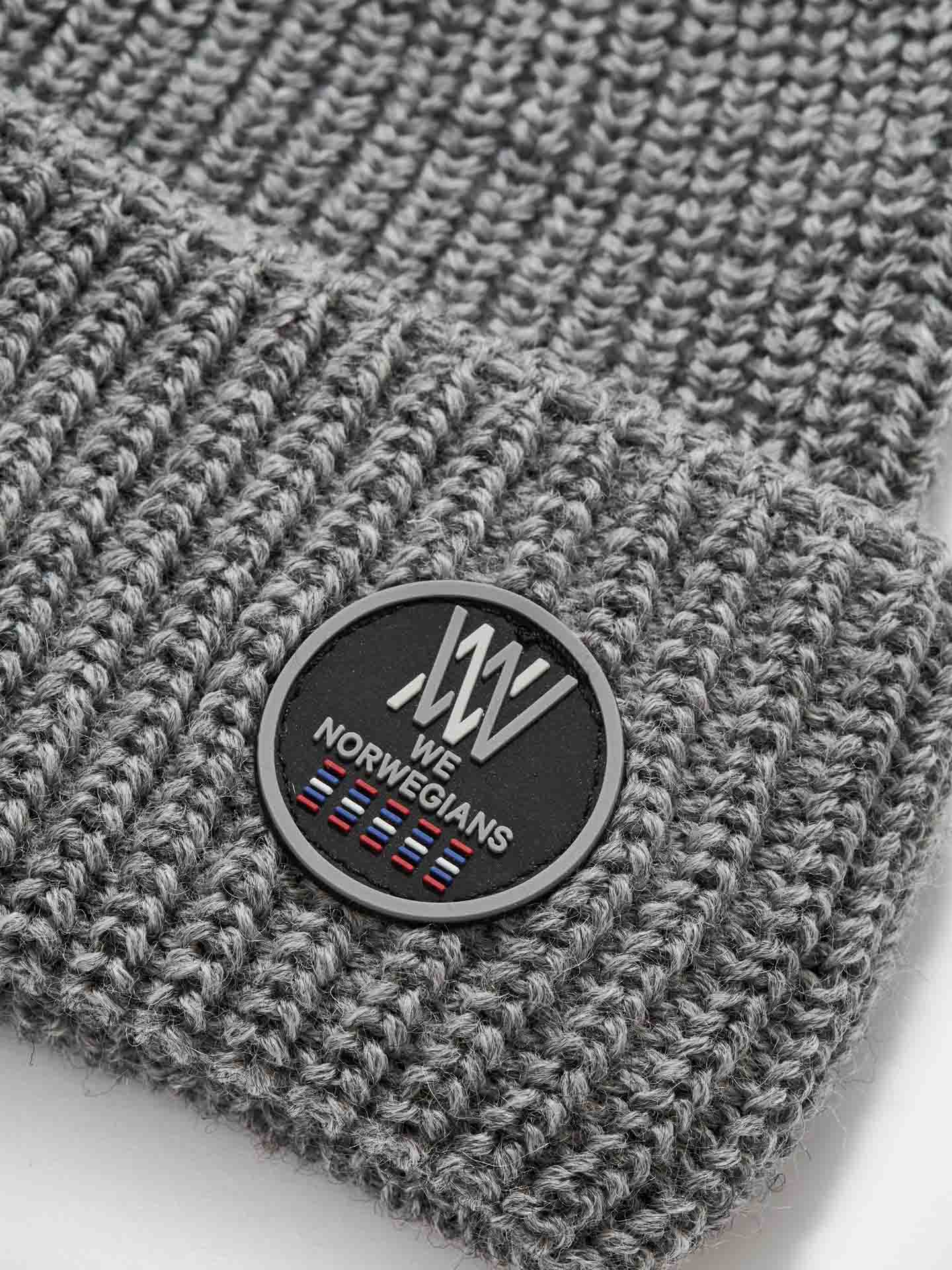 Close-up of the Kvitfjell Hat, a gray knitted beanie from We Norwegians, featuring a black circular patch displaying the brand logo, the text "We Norwegians," and a row of small flags at the bottom. Made from breathable Merino wool, the beanie's texture is prominently ribbed.