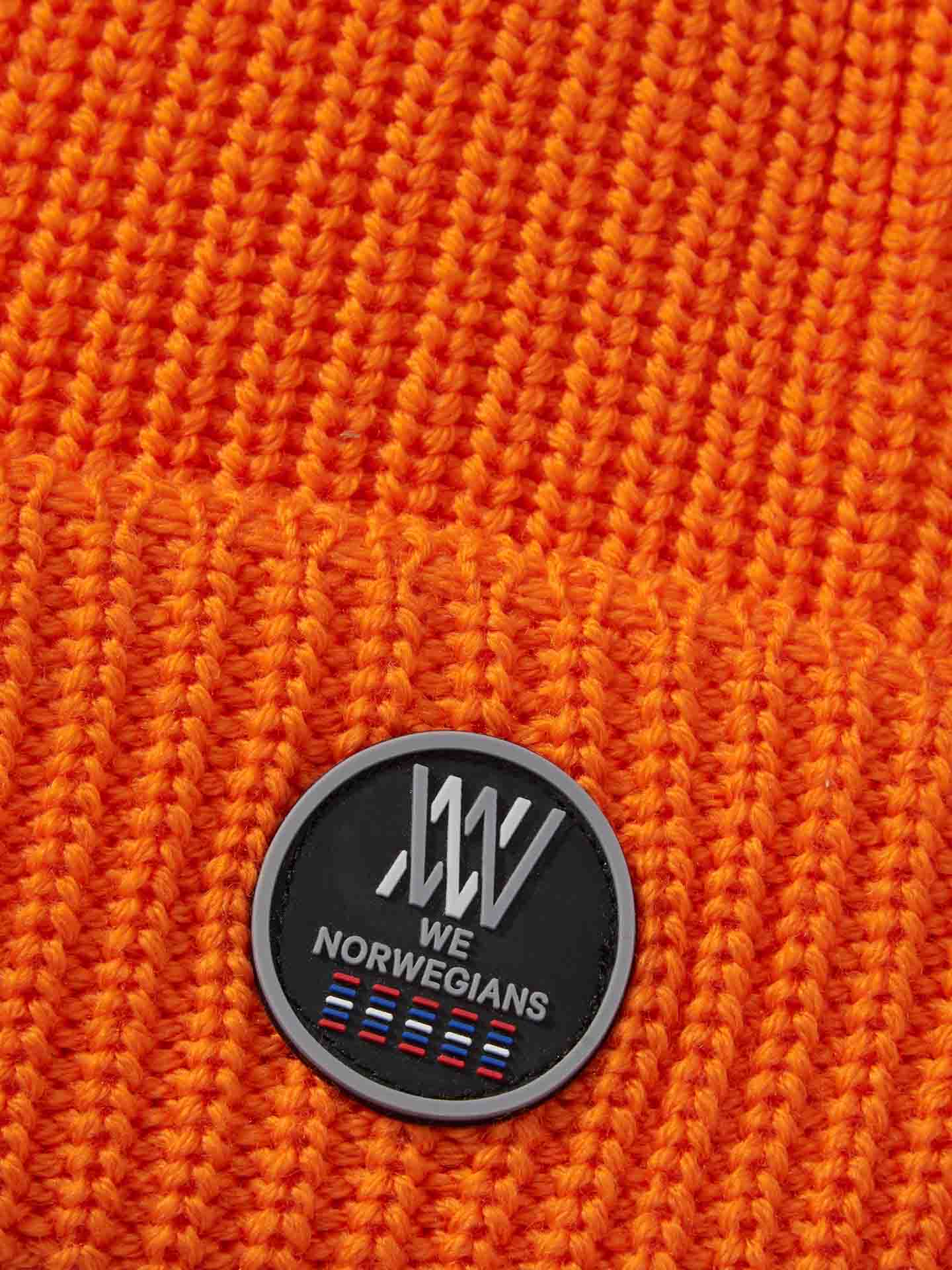 Close-up of an orange Kvitfjell Hat by We Norwegians, featuring a round black patch with stylized white text reading "We Norwegians." Below the text are small colored stripes representing various flags. The knitted texture and stitch patterns of the breathable, antibacterial beanie are clearly visible.