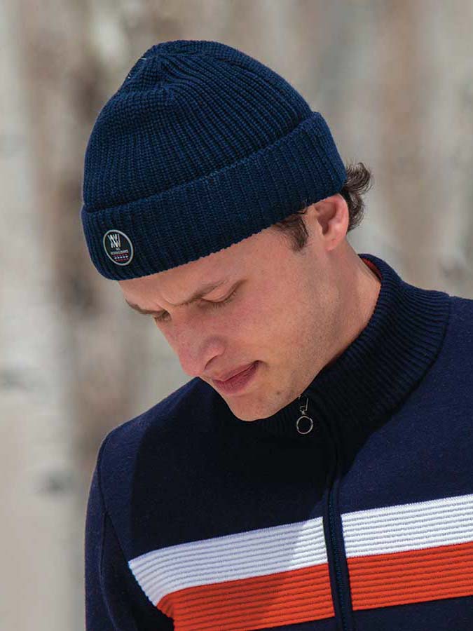 A man wearing the We Norwegians Kvitfjell Hat and a navy blue zip-up sweater with white and red stripes, crafted from breathable Merino wool, looks down pensively. The background is blurred, suggesting an outdoor setting.