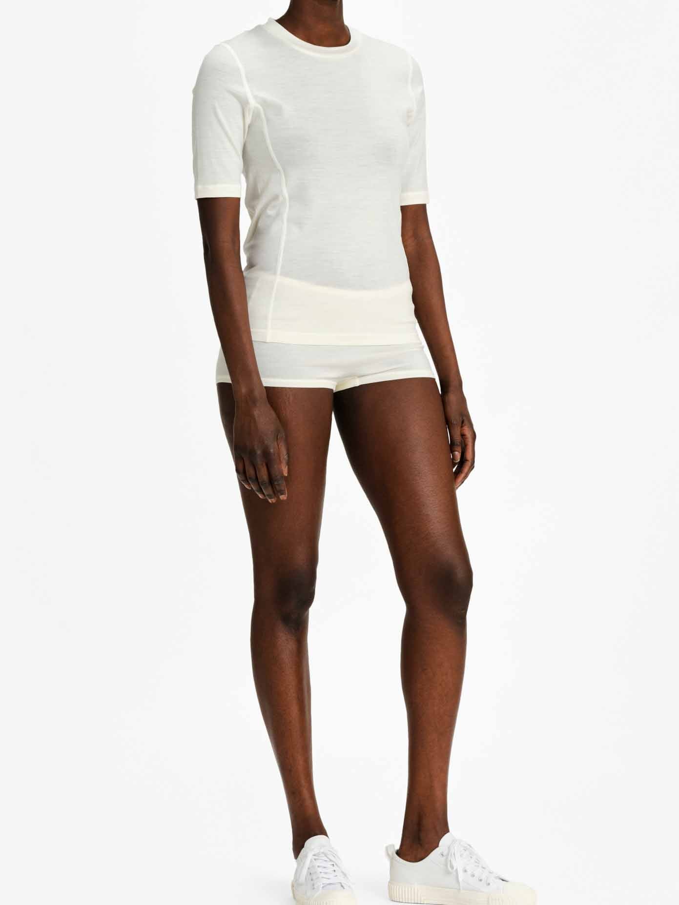 A person is dressed in the Sno Merino Base Layer Tee Women Off White from We Norwegians, along with matching shorts and odor-resistant white sneakers. They are standing against a plain white background.