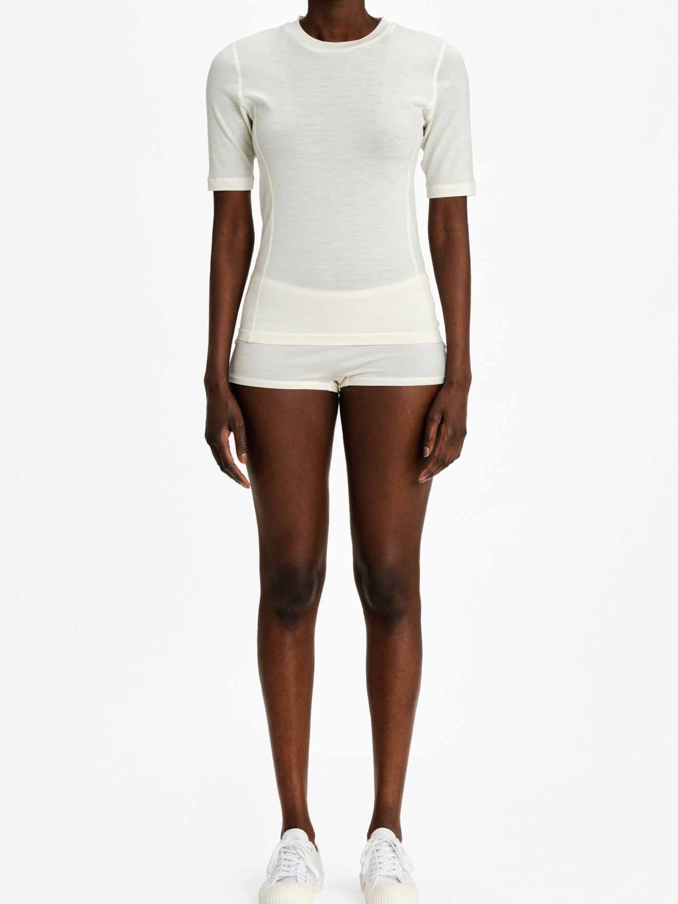 A person stands against a plain background wearing an off-white Sno Merino Base Layer Tee by We Norwegians and matching shorts. They are also wearing white sneakers. Their arms are relaxed by their sides, and their stance is upright.