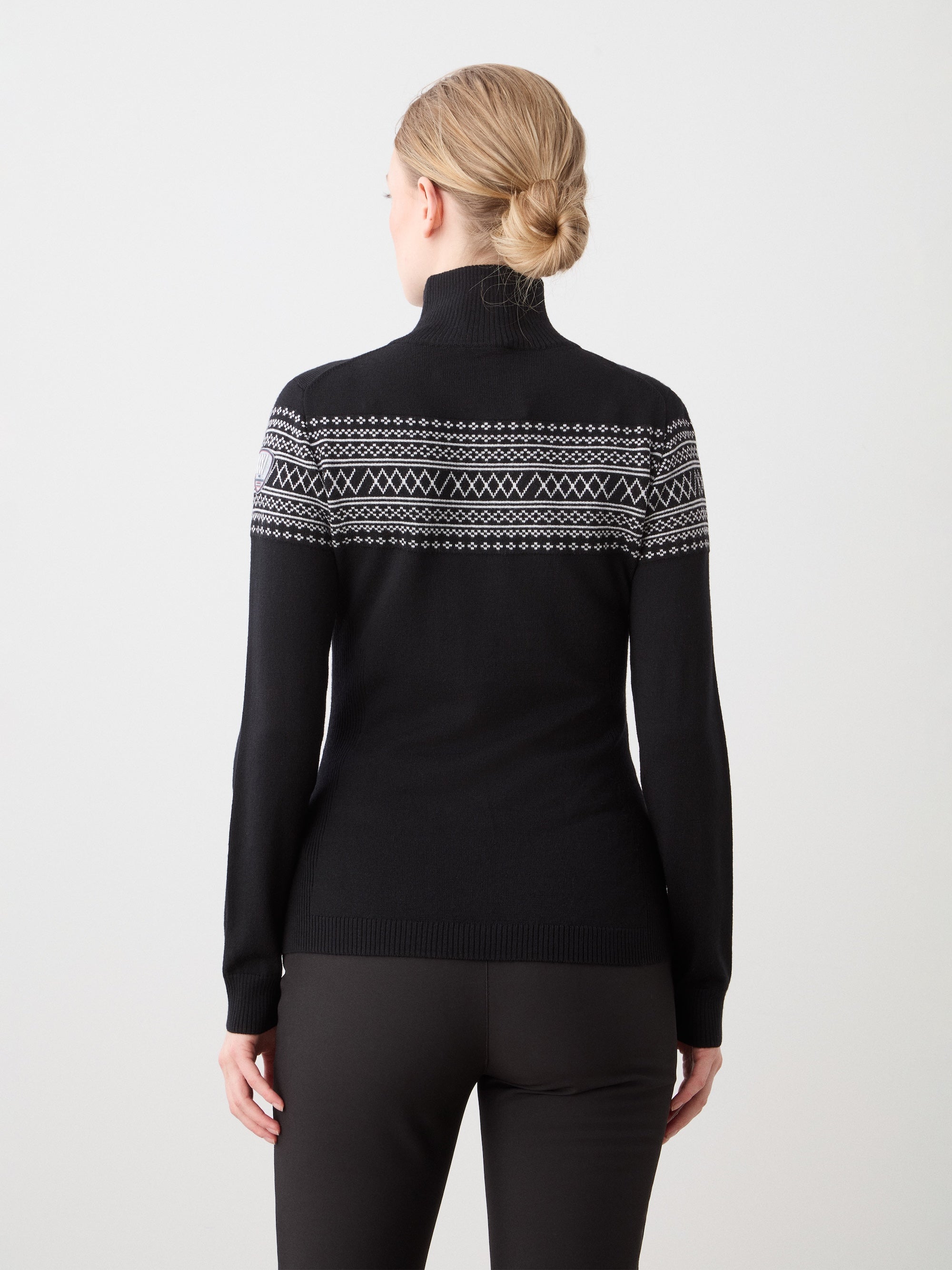 A woman with blonde hair in a bun is seen from behind wearing the We Norwegians Signature Zip Up Women, a black sweater with white Setesdal pattern, paired with dark pants, standing against a plain white background.