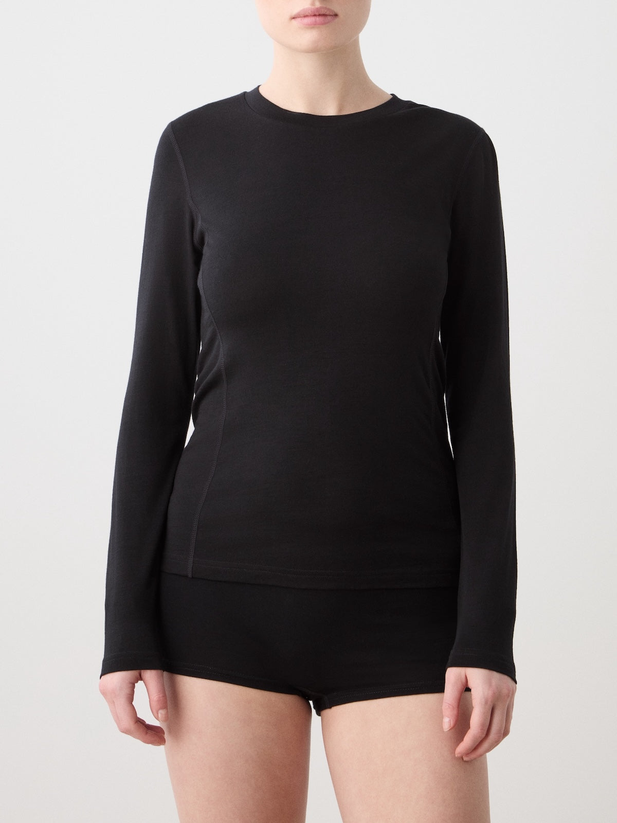 A person stands against a plain light background wearing the We Norwegians Sno Merino Crewneck Women—a fitted black long-sleeve shirt paired with black shorts, both crafted from breathable single jersey. Their face is partially visible and their arms are relaxed at their sides.