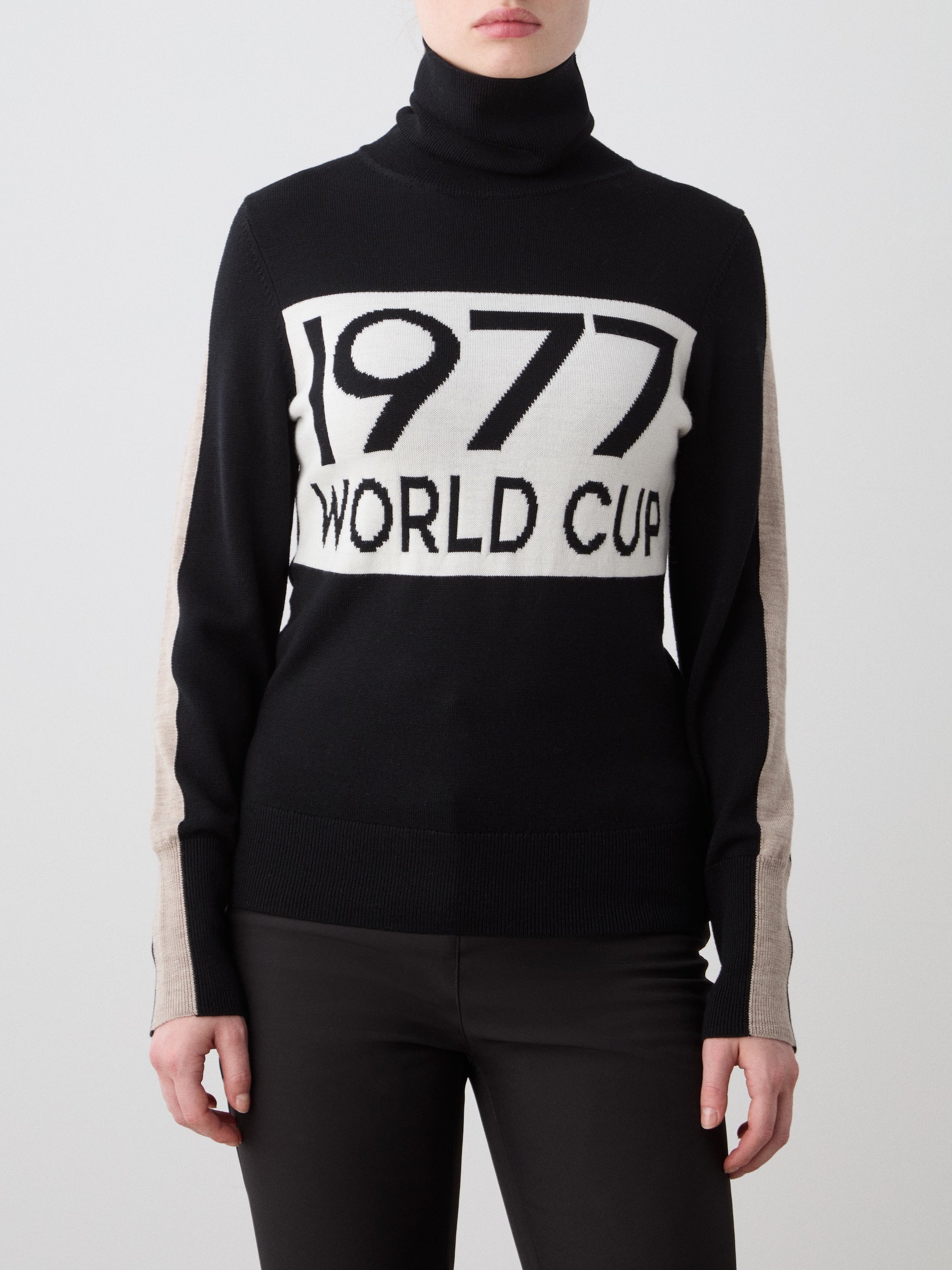 A woman wears the We Norwegians 1977 World Cup Sweater, featuring a black 70's ski-inspired style with "1977 WORLD CUP" on a white front panel, beige sleeve stripes, and pairs it with black pants.