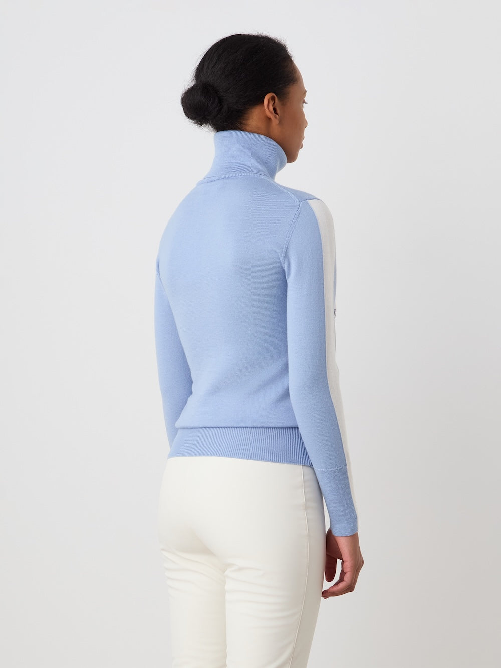 A woman with dark hair in a bun wears the We Norwegians World Cup Sweater women - Pre Spring 26 (light blue merino wool) and white pants, standing with her back to the camera against a plain white background.