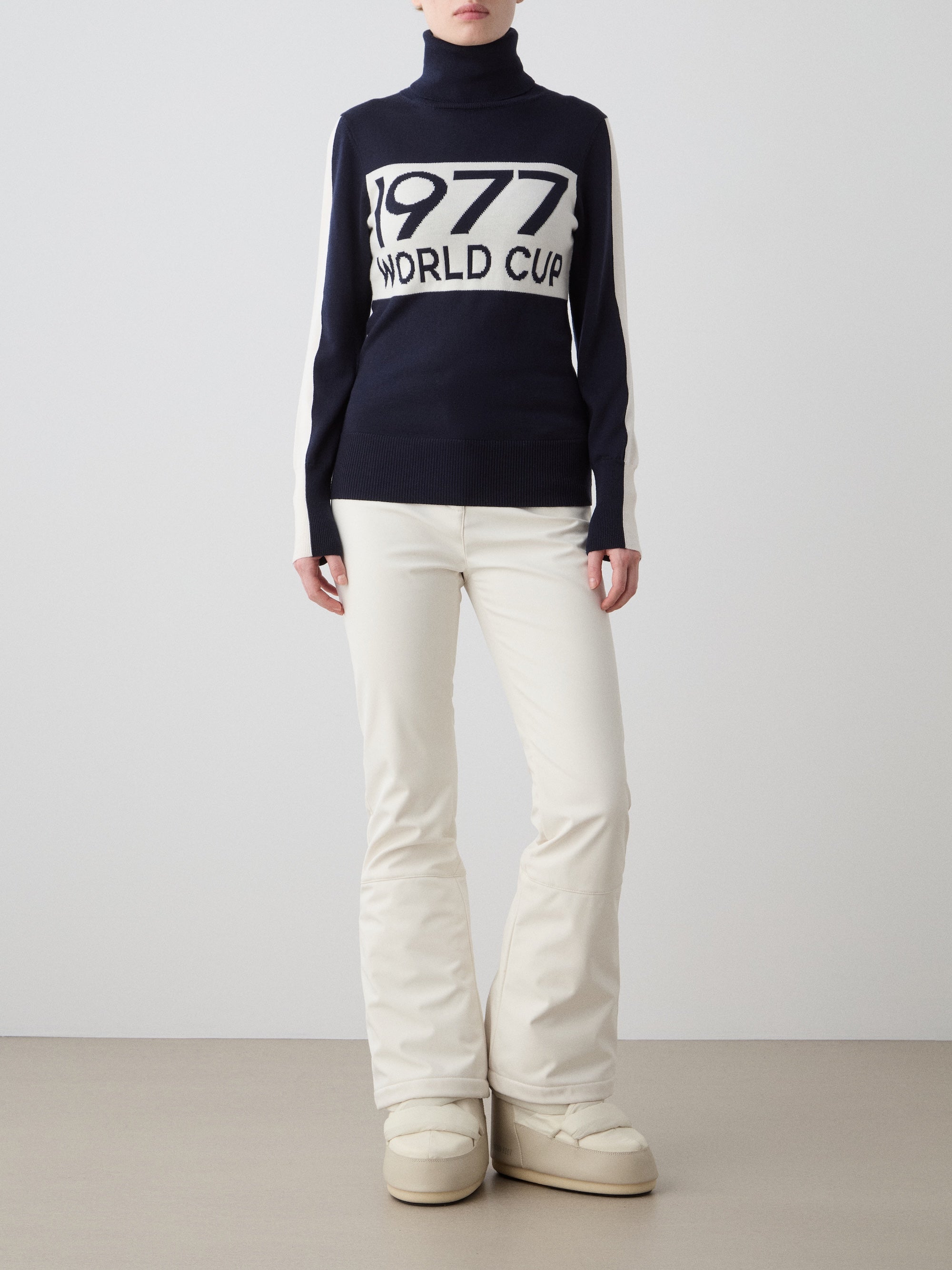 A woman wears the We Norwegians 1977 World Cup Sweater, featuring "1977 WORLD CUP" on navy, paired with white pants and cream snow boots for a chic retro ski look against a plain background.