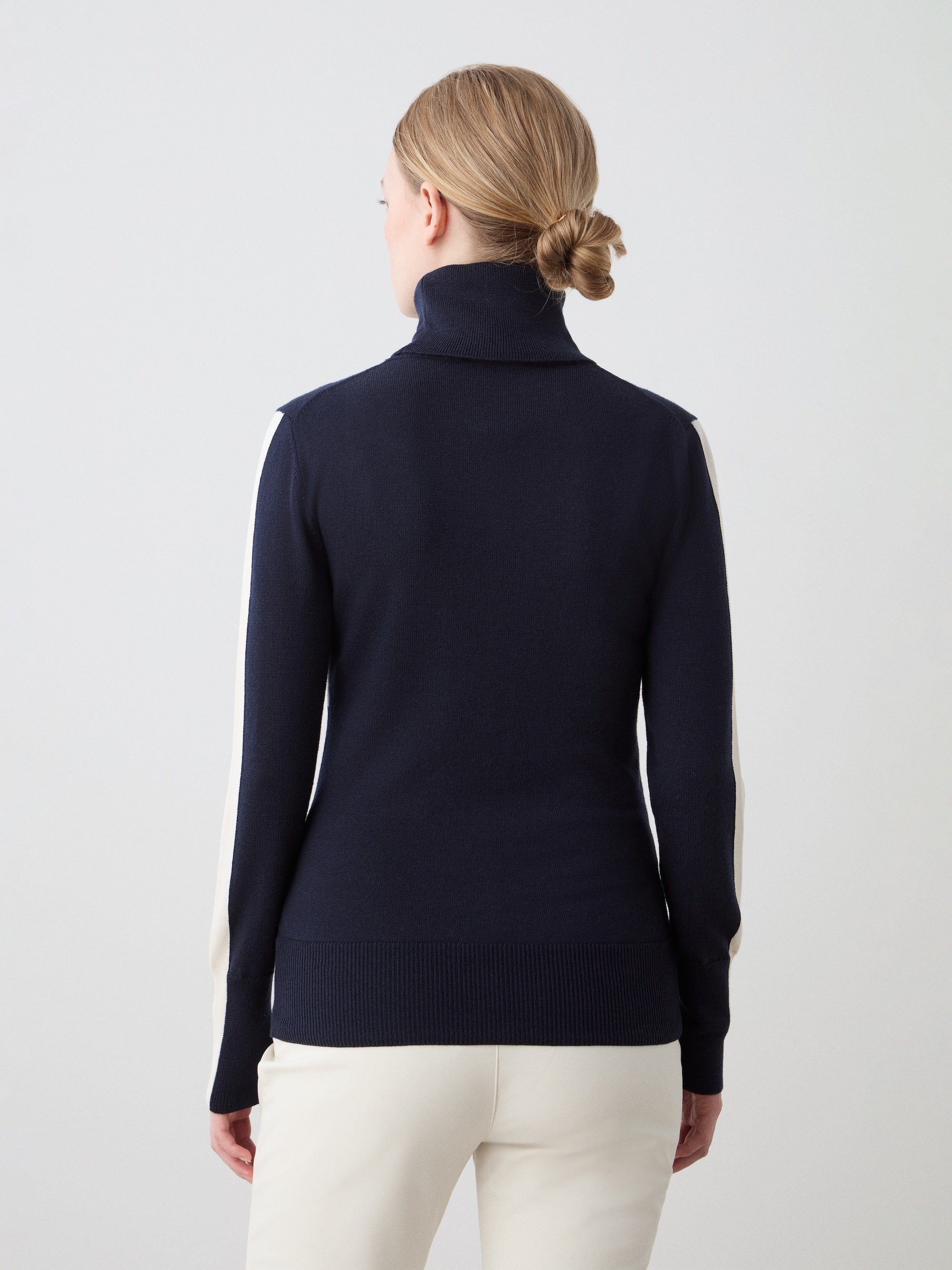 A woman with blonde hair in a low bun is seen from behind, wearing the We Norwegians 1977 World Cup Sweater Women with white sleeve stripes and off-white pants, channeling classic 70’s ski style against a simple light background.