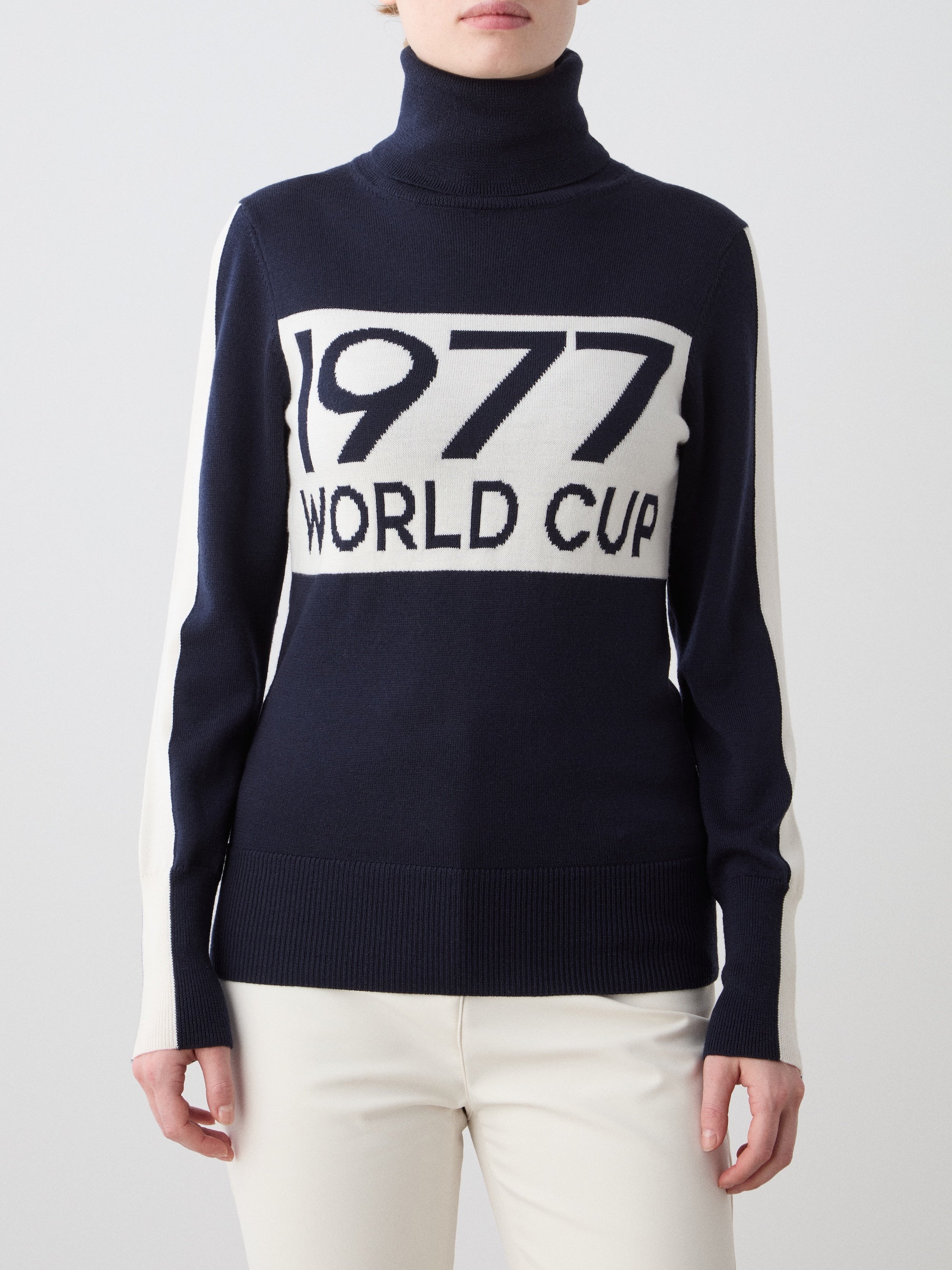 A woman wears the We Norwegians 1977 World Cup Sweater in navy with white sleeve and front panels printed with "1977 WORLD CUP," styled with crisp white pants for a classic ‘70s ski look.