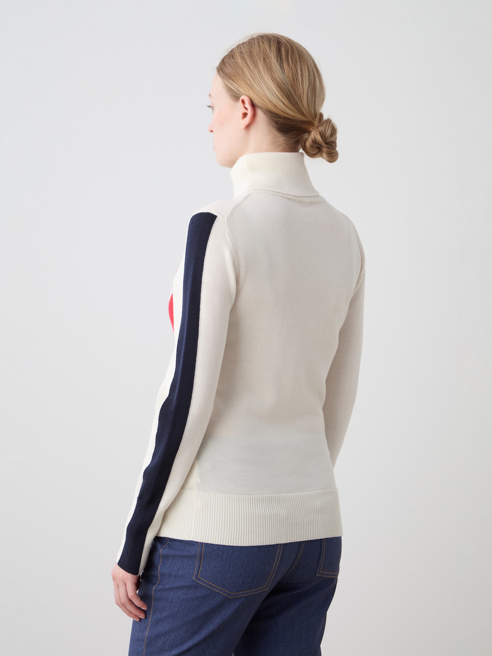 A woman with blonde hair in a low bun models the We Norwegians 1977 World Cup Sweater Women—cream with dark sleeve stripes—paired with blue jeans, evoking classic '70s ski style as she stands with her back to the camera against a plain background.