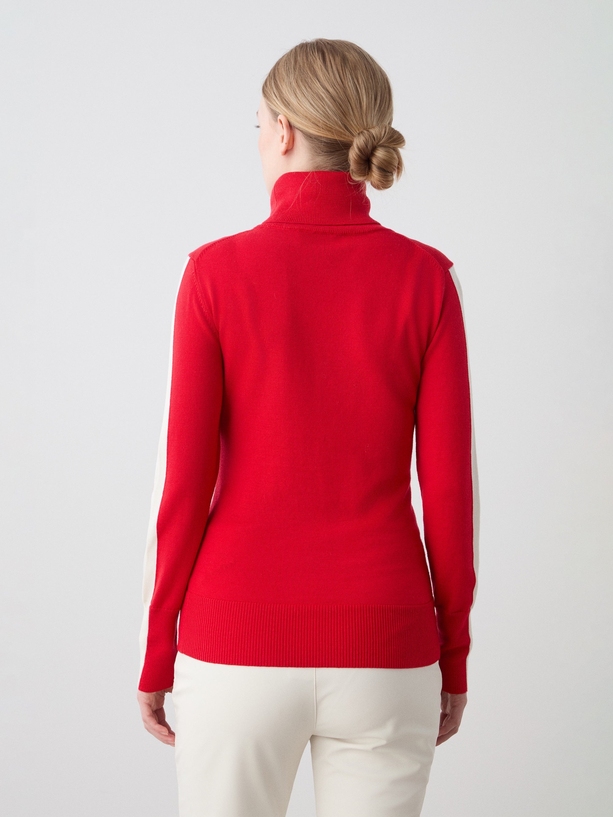 A woman with blonde hair in a neat bun, seen from behind, wears the We Norwegians 1977 World Cup Sweater Women—a red ski sweater with white sleeve stripes—and white pants, evoking classic ‘70s skiing style against a plain light background.