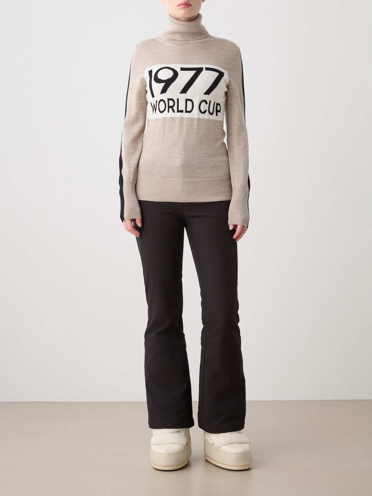 A woman models the We Norwegians 1977 World Cup Sweater in beige merino wool, paired with black flared pants and white boots, standing against a plain background and capturing the essence of retro ski fashion.