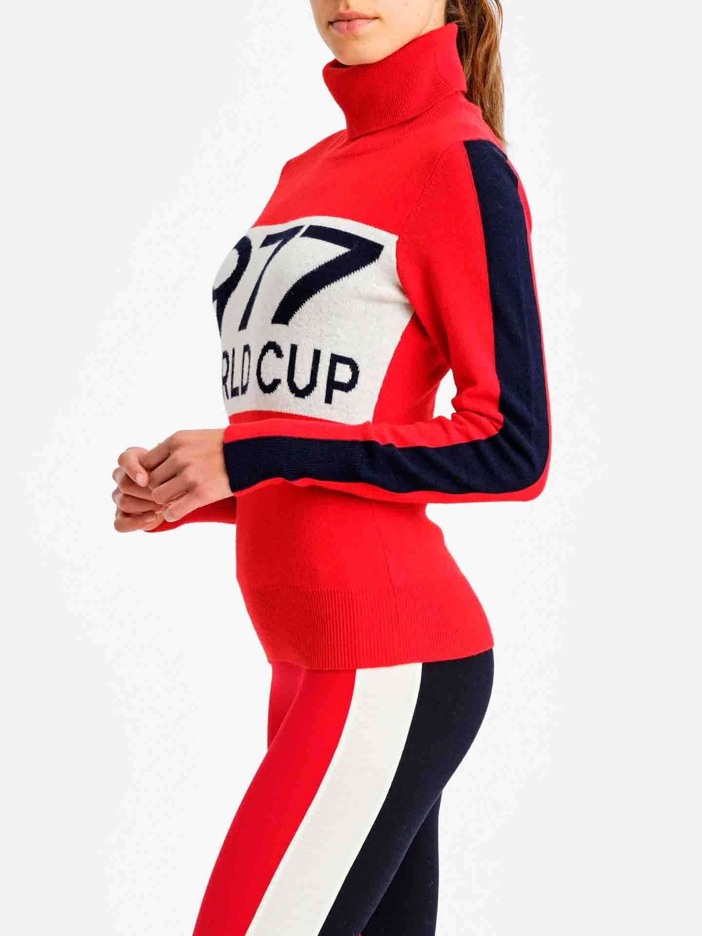A person wearing a We Norwegians' 1977 World Cup Sweater in red, adorned with "1977 WORLD CUP" on the chest. Reminiscent of classic ski fashion, the sweater features a white panel on the front and navy blue stripes along the sleeves. They complete their look with matching red and navy blue leggings that have crisp white side stripes.