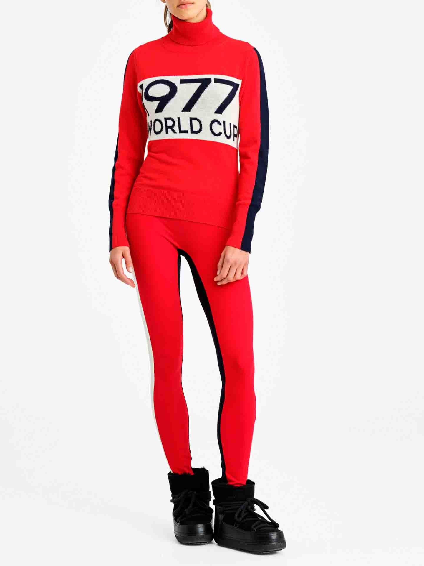 A person is wearing the We Norwegians 1977 World Cup Sweater Women Red, which has "1977 WORLD CUP" written on the front. They are also wearing matching red leggings with black side panels and black ankle boots with a fuzzy trim. The person, embodying retro ski fashion, is standing against a plain white background.