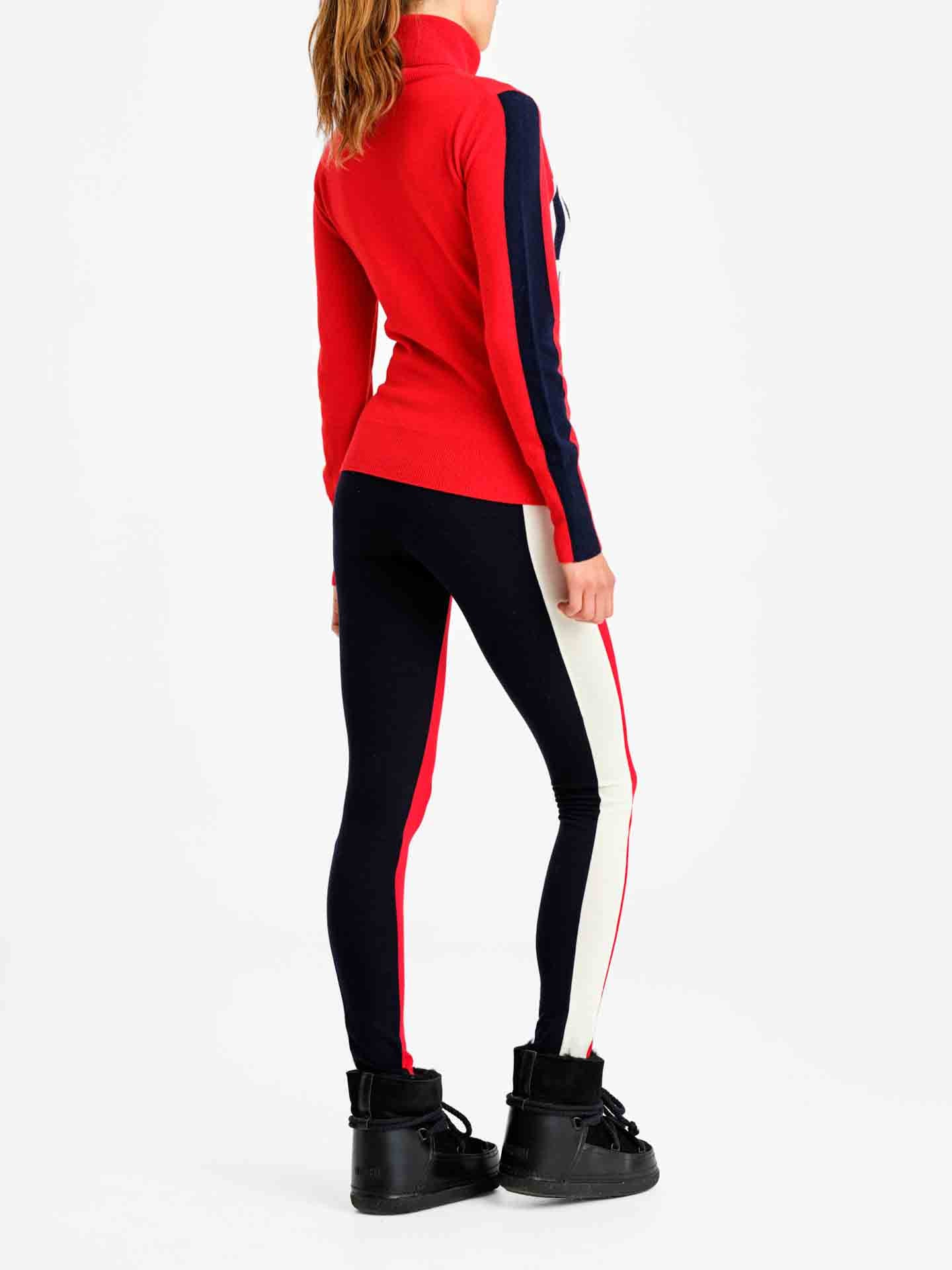 A woman stands with her back to the camera, showcasing a retro ski fashion. She is wearing the 1977 World Cup Sweater Women Red by We Norwegians, characterized by dark blue and white stripes on the sleeves. Matching dark blue leggings feature red and white side stripes, complemented by black snow boots. The background is plain white.