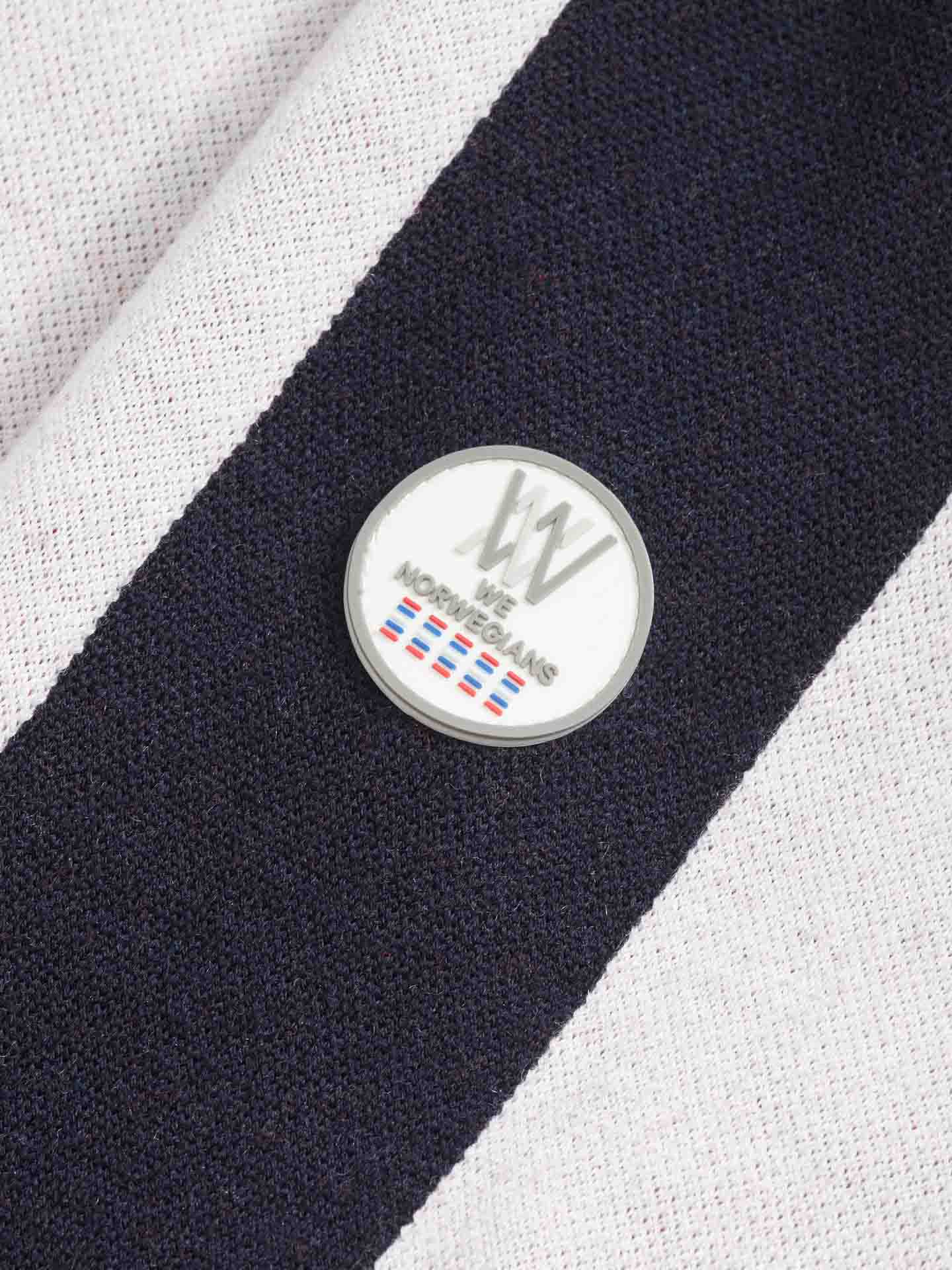 A close-up image showcases the fine merino wool fabric of the Hafjell Crewneck Women White by We Norwegians, featuring black and white stripes. Attached to the fabric is a round button displaying the "WW" logo along with white text that reads "We Norwegians." The button also includes small multicolored horizontal lines.