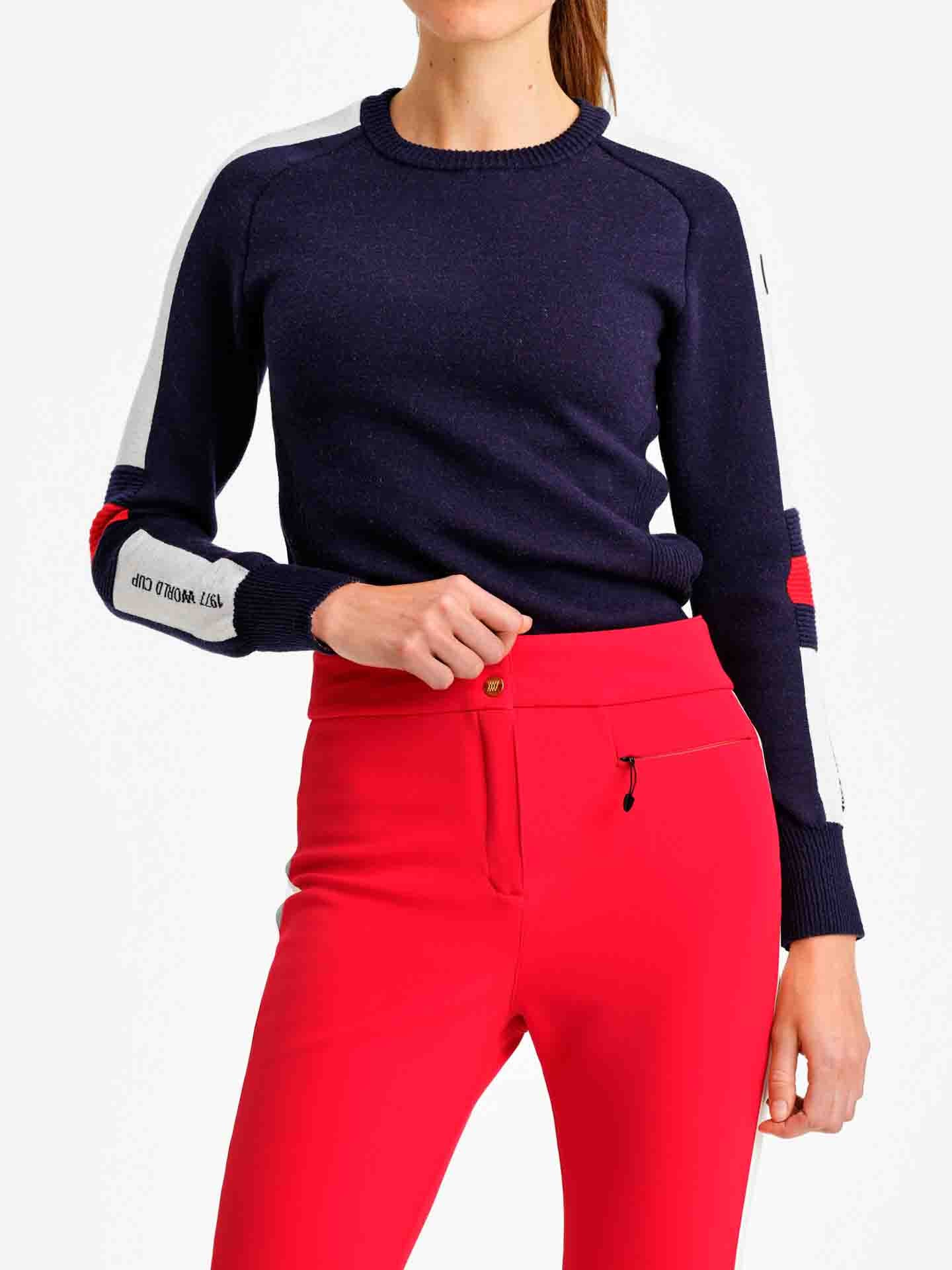A person is wearing the Hafjell Crewneck Women Flag sweater by We Norwegians, featuring a navy blue color with fine merino wool and white and red accents on the sleeves, paired with bright red pants. The person has their left hand resting lightly on their hip against a plain white background.