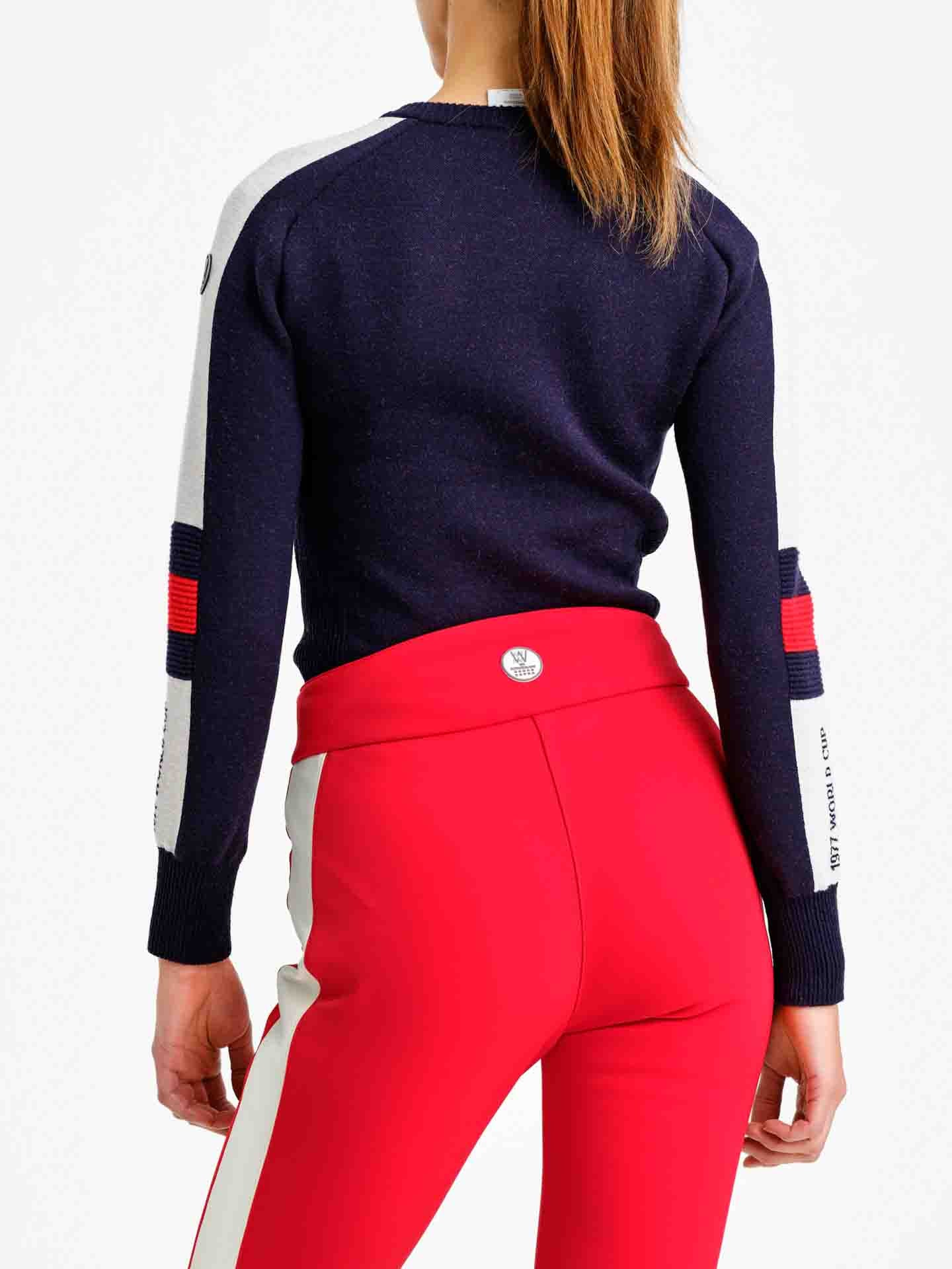 A person is standing with their back to the camera, showcasing a We Norwegians Hafjell Crewneck Women Flag ski sweater made of fine merino wool. The navy blue sweater features white and red accents on the sleeves, paired with fitted red pants that have a white stripe down the sides. Their hair is tied back in a ponytail.