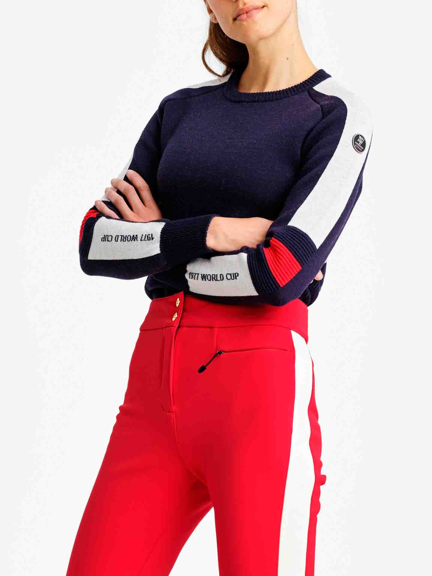 A woman crosses her arms, donning the Hafjell Crewneck Women Flag sweater by We Norwegians, adorned with white and red stripes on its navy blue sleeves and finely crafted from merino wool. The text on the sleeves reads "FIS Ski World Cup" and "Audi FIS Ski World Cup." She pairs the stylish sweater with high-waisted red pants accented by white side stripes.