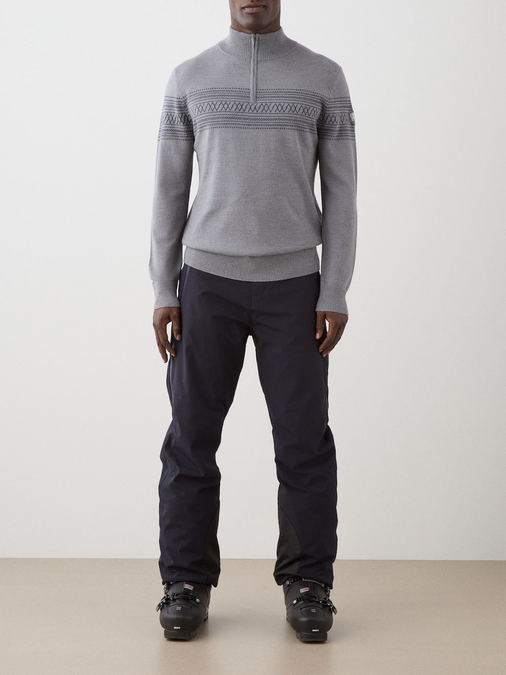 A person wearing the We Norwegians Signature Zip Up Men, a light gray sweater featuring a classic Norwegian Setesdal pattern, along with dark pants and black ski boots, stands facing forward against a plain background.