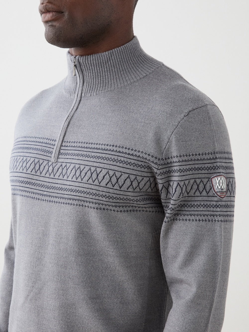 A person wears the We Norwegians Signature Zip Up Men, a light gray merino wool sweater featuring a collared zip-up design and dark geometric Setesdal-inspired patterns across the chest and arms. The left sleeve is adorned with a small patch displaying roman numerals, and the sweater is crafted from washable merino wool for easy care.