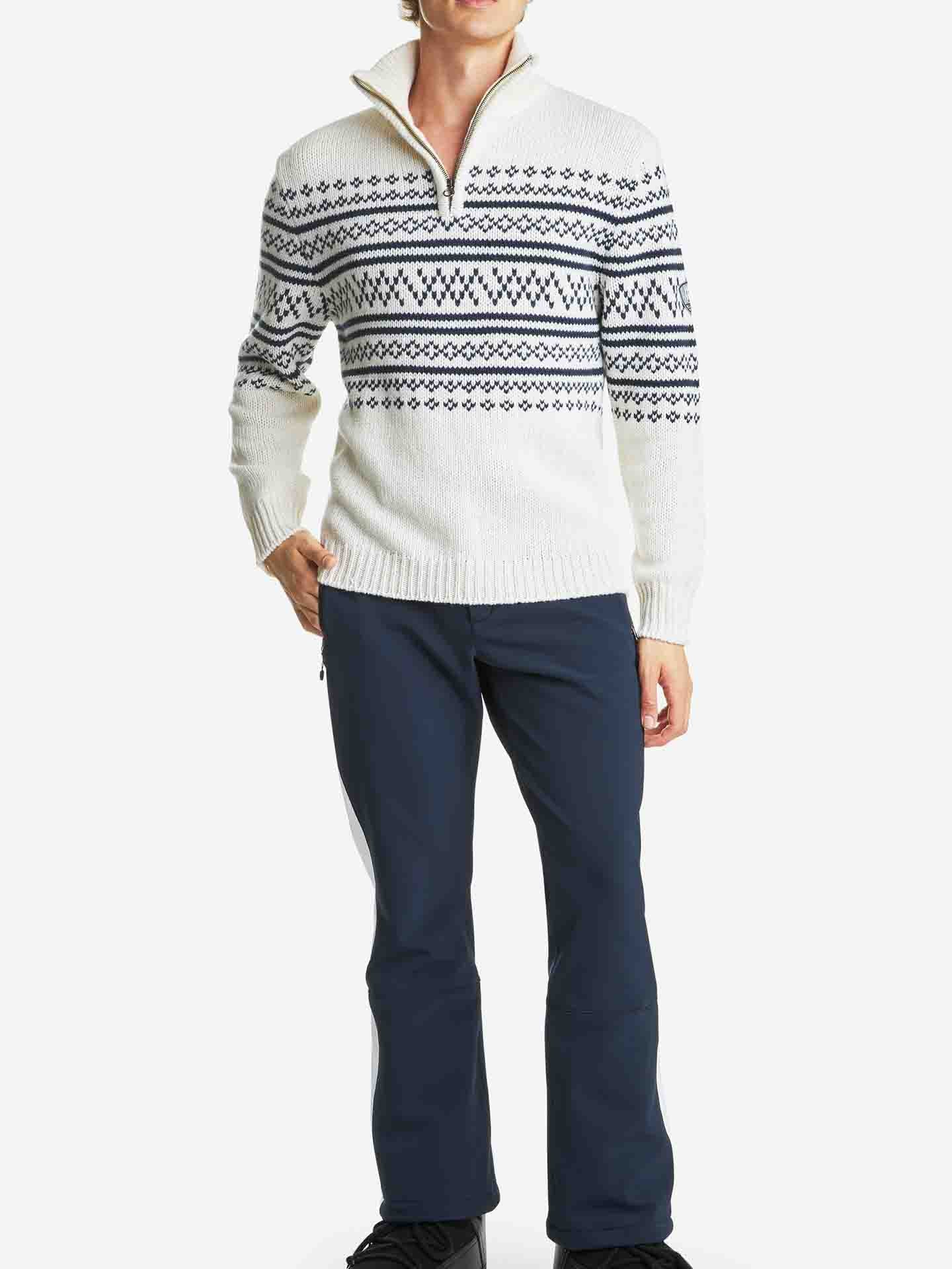 A person is wearing the "Setesdal Zip Up Sweater Men Off White" by We Norwegians, featuring a black diamond pattern across the chest and sleeves, paired with navy blue pants and black shoes. The chunky knits give an après ski vibe as the individual stands with their right hand in their pants pocket against a plain white background.
