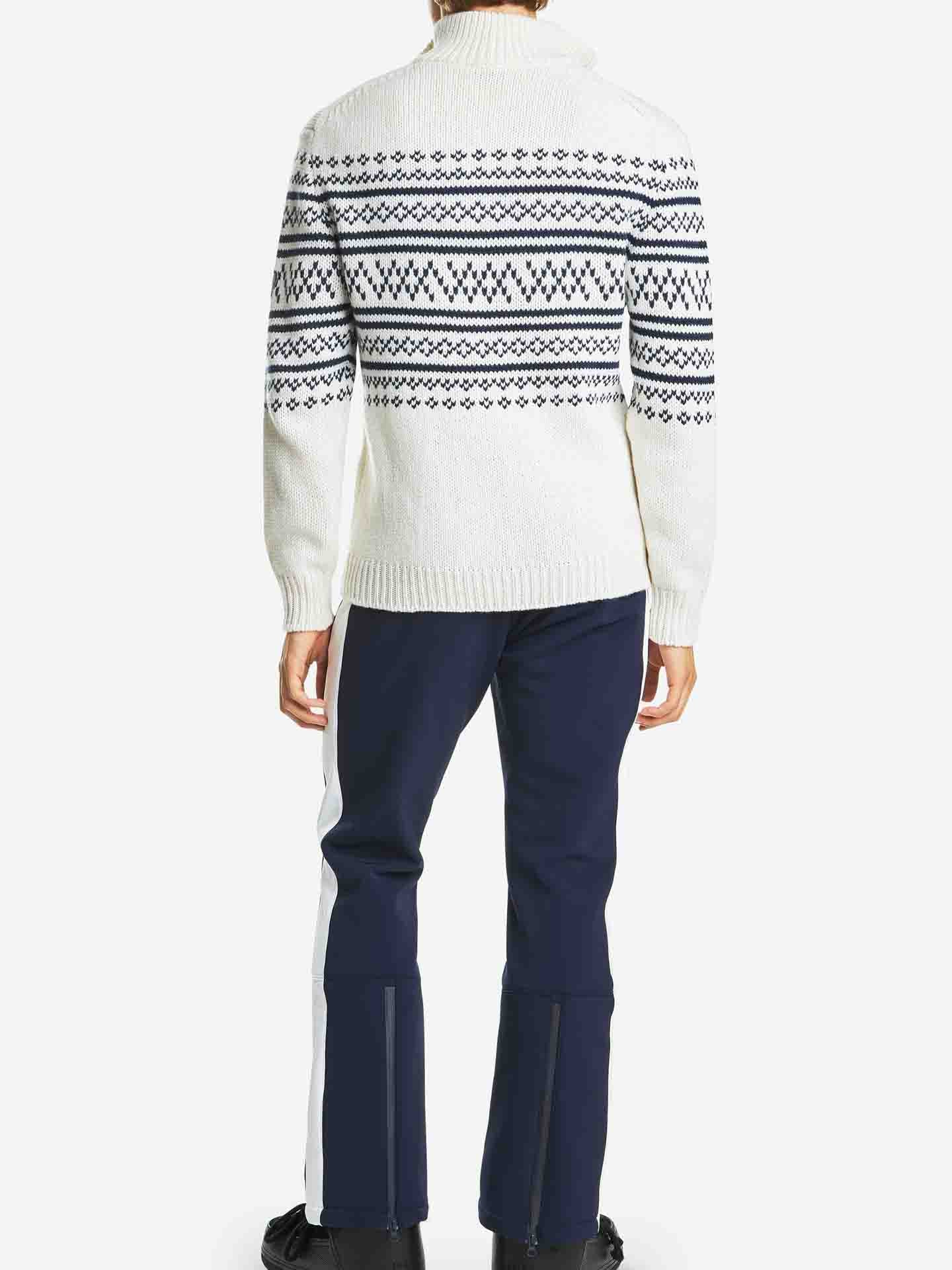 A person wearing the We Norwegians Setesdal Zip Up Sweater Men Off White, characterized by its chunky knit design and black and blue geometric patterns on the back, along with dark blue pants featuring white stripes on the sides, is standing facing away from the camera. The background is plain white.